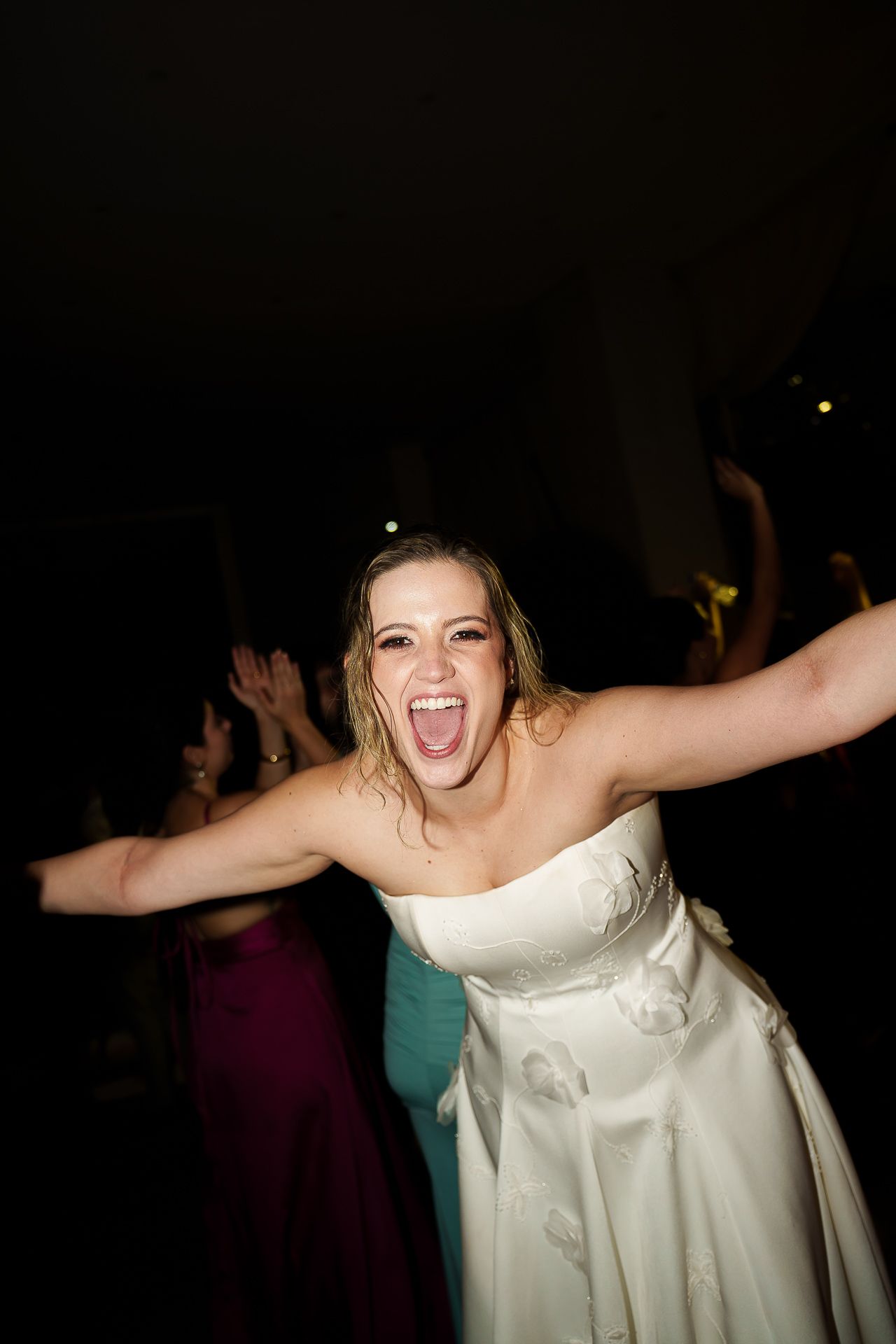 Joyful bride in white strapless gown dancing with arms outstretched at wedding reception.