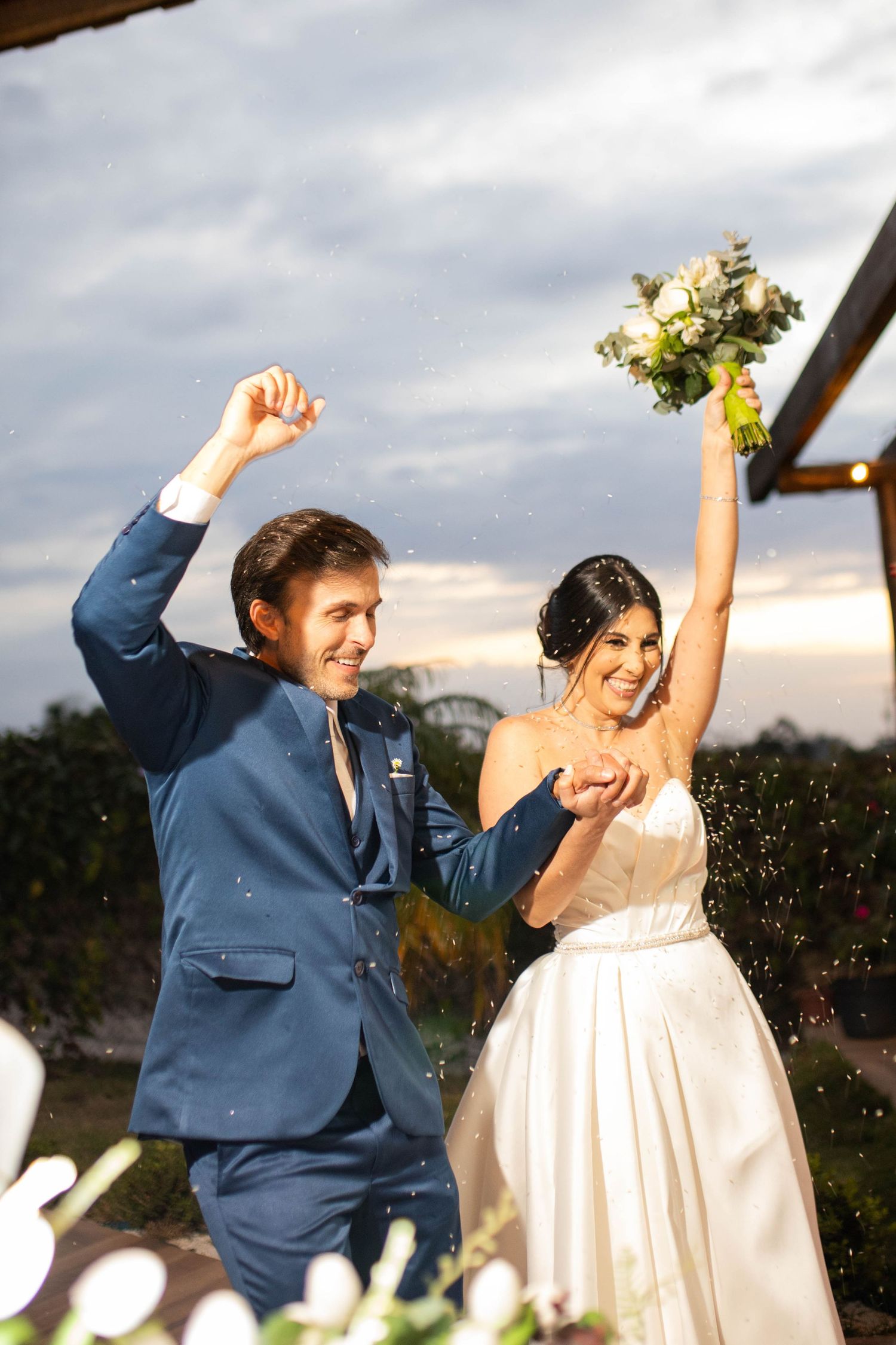 Joyful newlyweds celebrate after wedding ceremony, bride holding bouquet aloft, groom in blue suit, confetti falling at dusk.