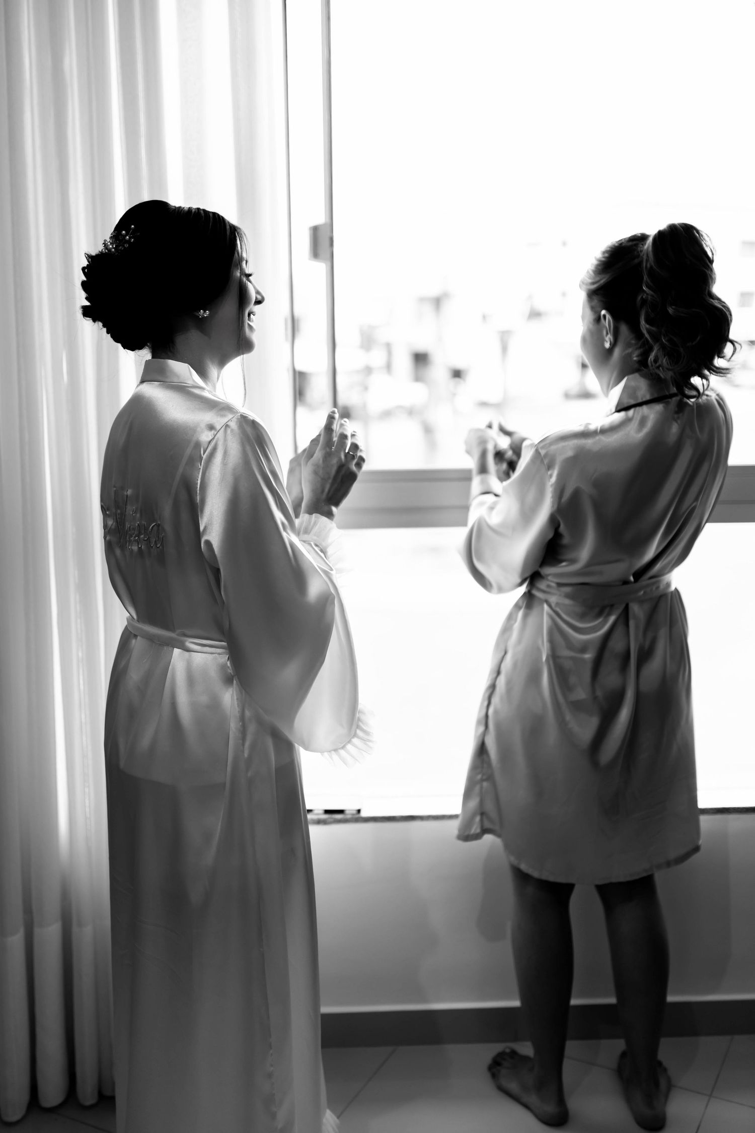 Two women in satin robes stand by a bright window, sharing a quiet moment before a wedding ceremony.