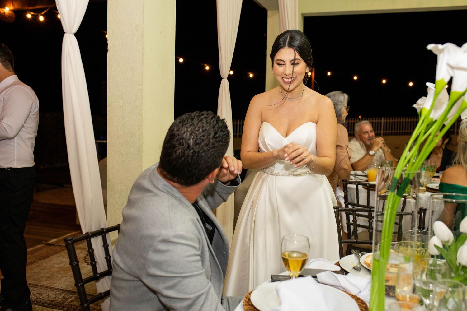 Bride in white satin gown laughing with seated guest at elegant outdoor wedding reception with floral centerpieces.