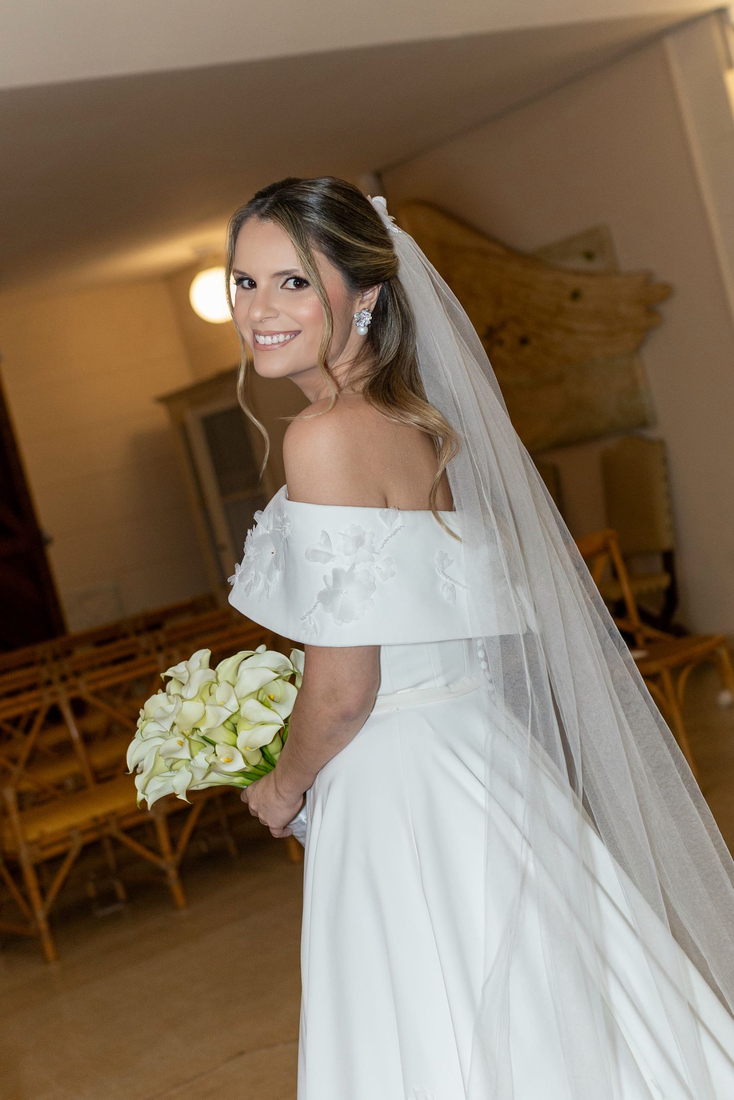 Elegant bride in off-shoulder wedding dress with veil, holding white calla lily bouquet, smiling.