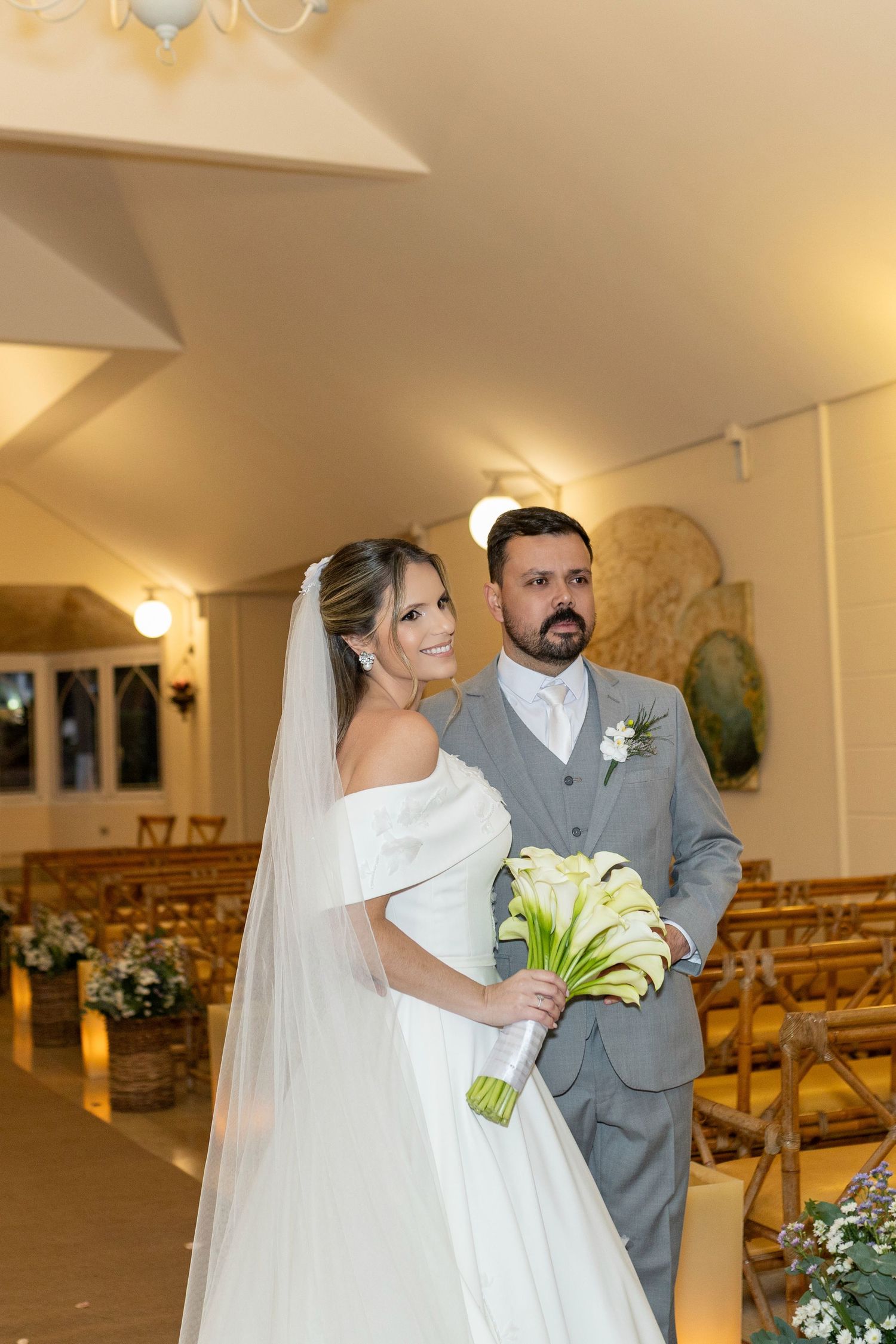 Bride in white off-shoulder gown with veil holds calla lily bouquet beside groom in gray suit in chapel aisle.