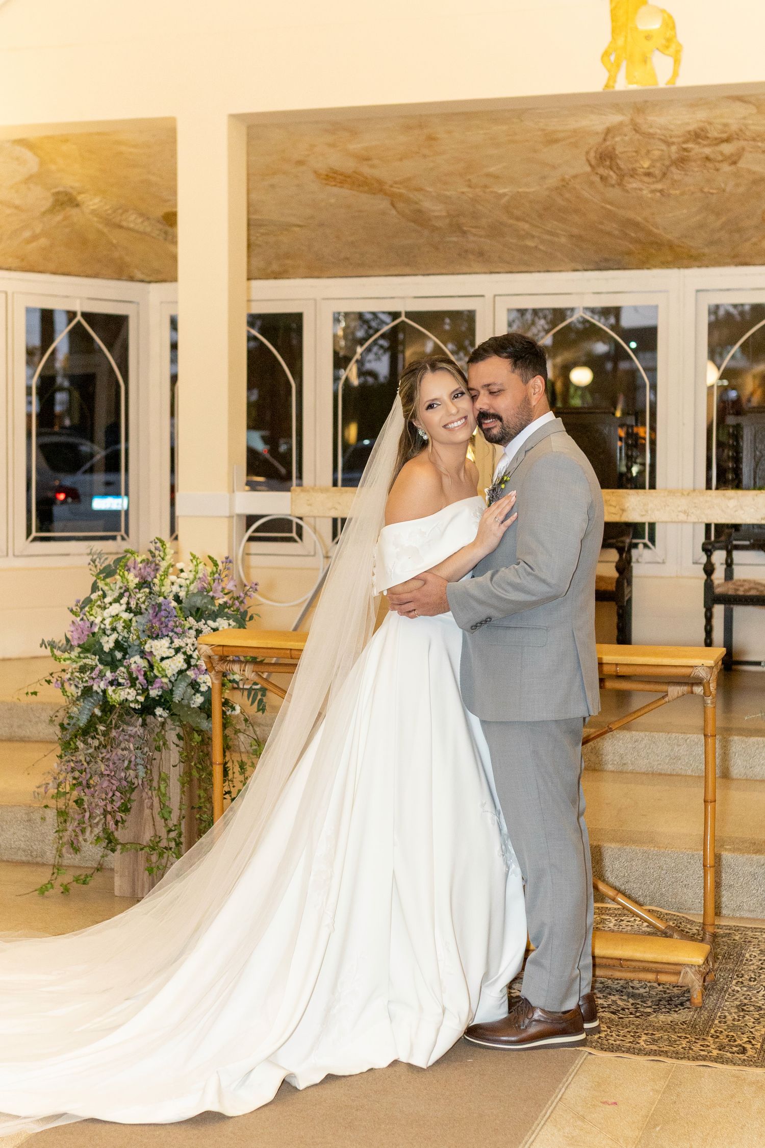 Bride in white gown with veil and groom in gray suit embrace at church altar with floral arrangements.