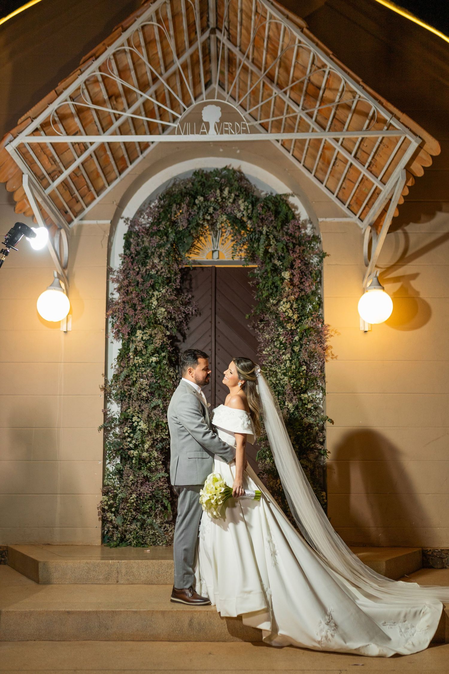 Bride and groom share romantic moment at floral-adorned chapel entrance with warm lighting at night.