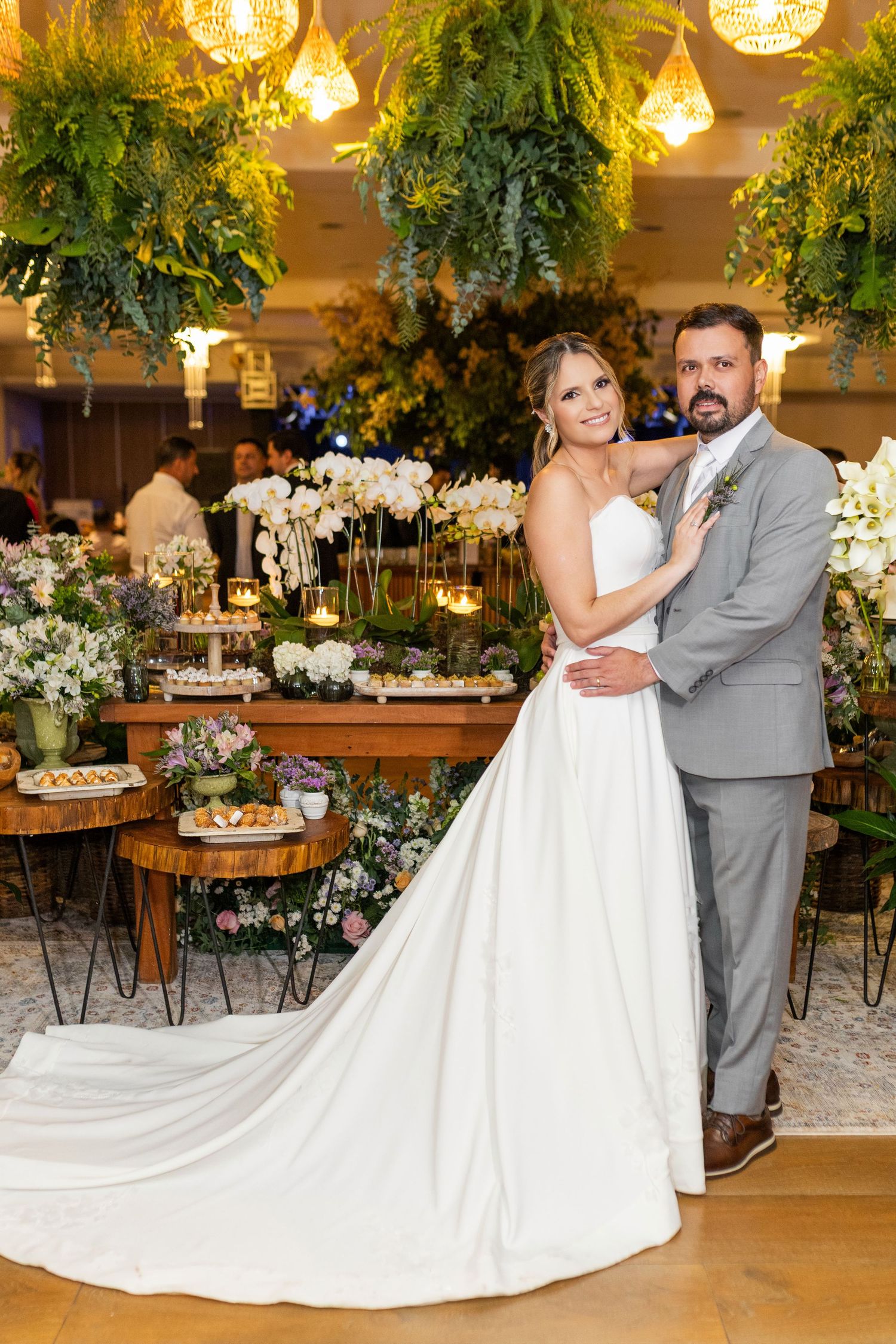 Bride in white gown and groom in gray suit pose at elegant wedding reception with floral decor and hanging greenery.