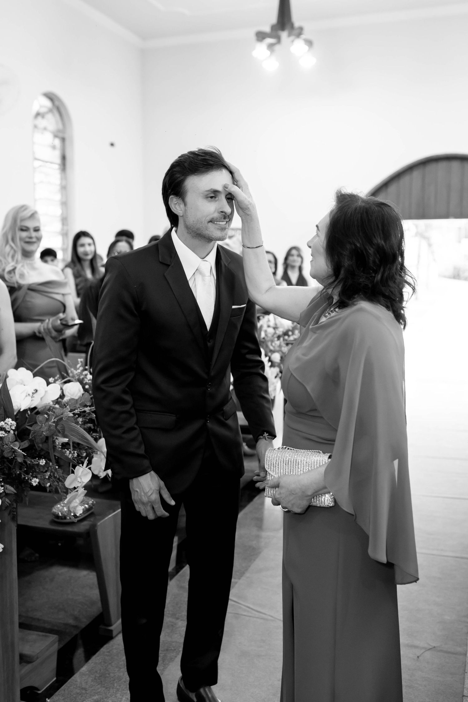 Black and white photo of a groom in a suit being touched on the face by a woman in a church wedding ceremony.