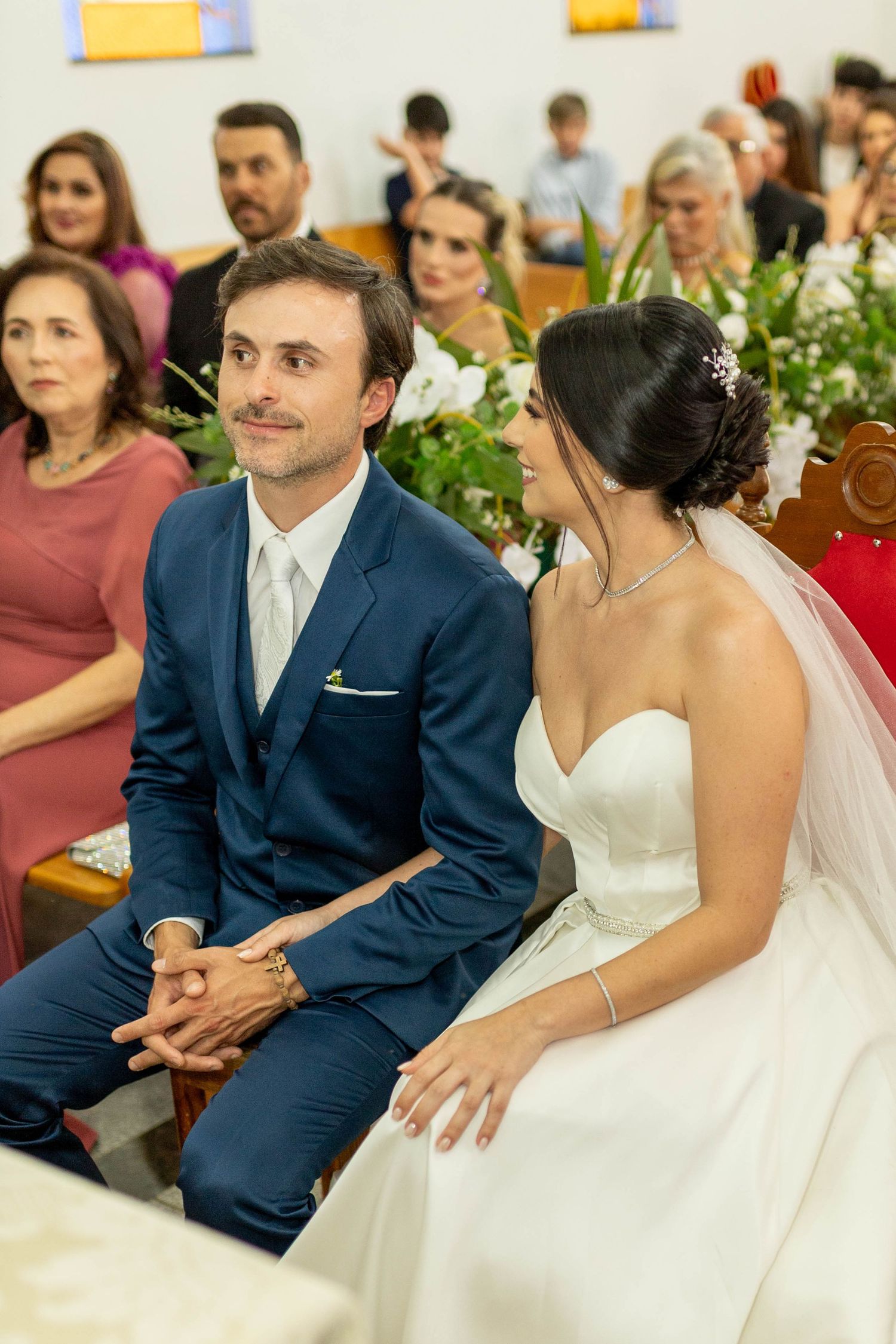 A bride and groom sit together at a church wedding ceremony, smiling at each other, surrounded by guests.