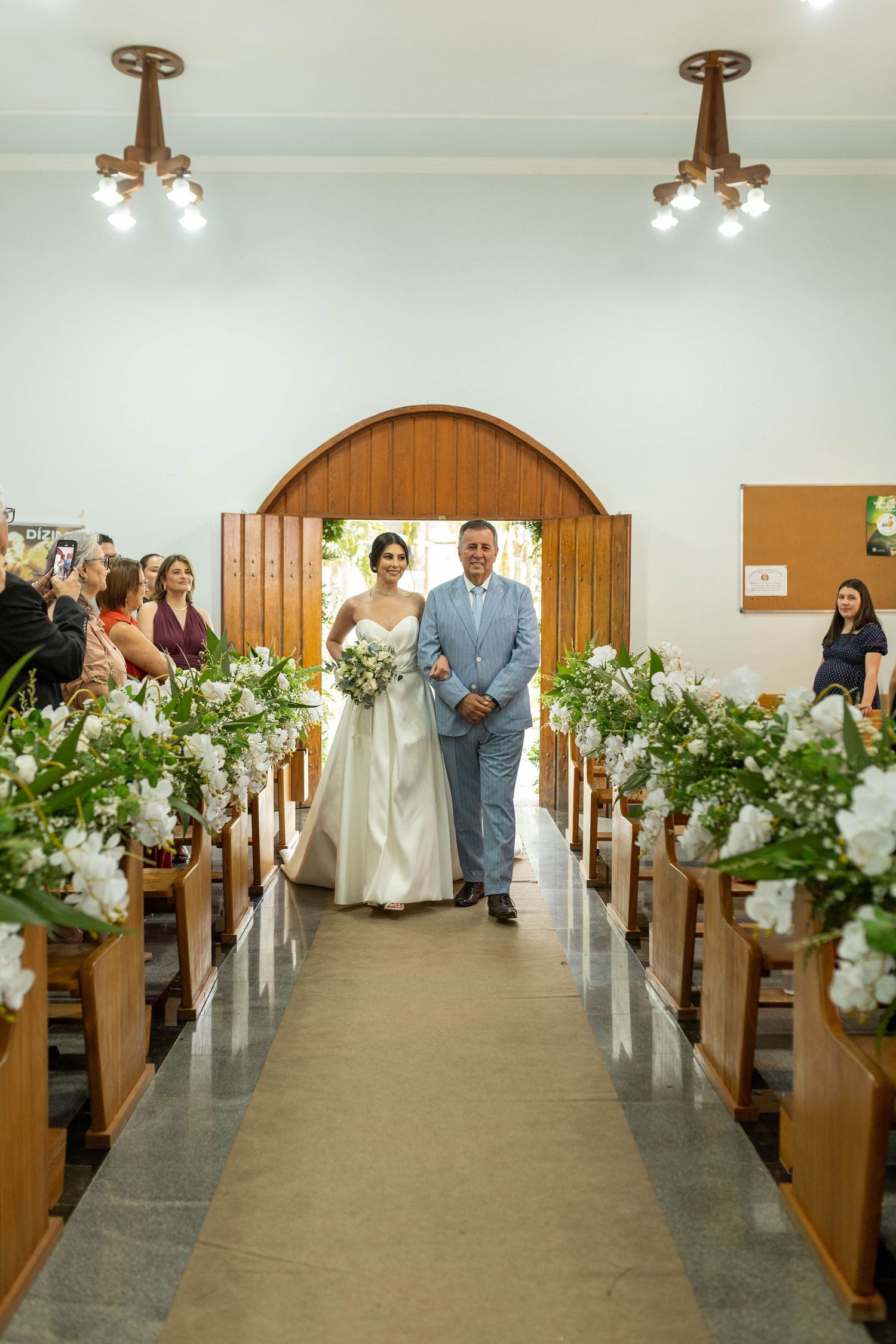 Bride walks down church aisle with escort, flanked by white floral arrangements on wooden pews during wedding ceremony.