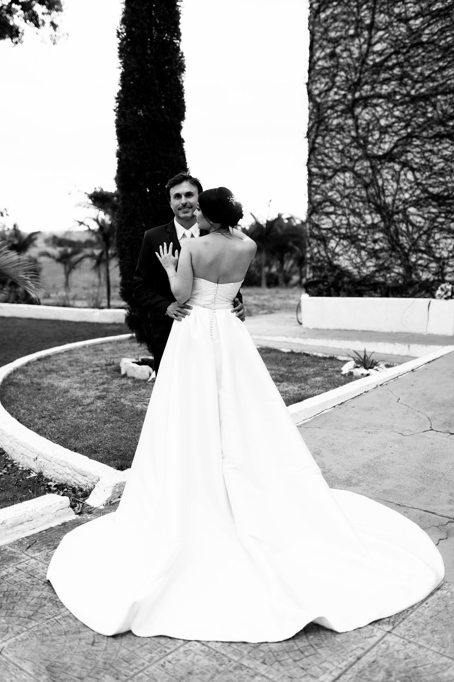 Black and white photo of bride and groom embracing outdoors, bride in strapless gown with long train.