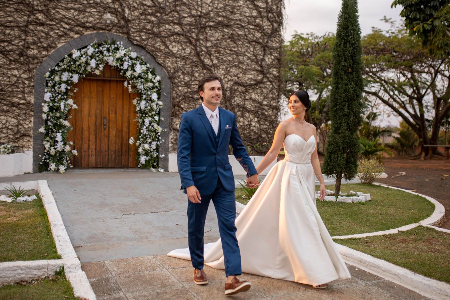 Bride and groom walk hand-in-hand outside a stone chapel adorned with white floral arch on their wedding day.