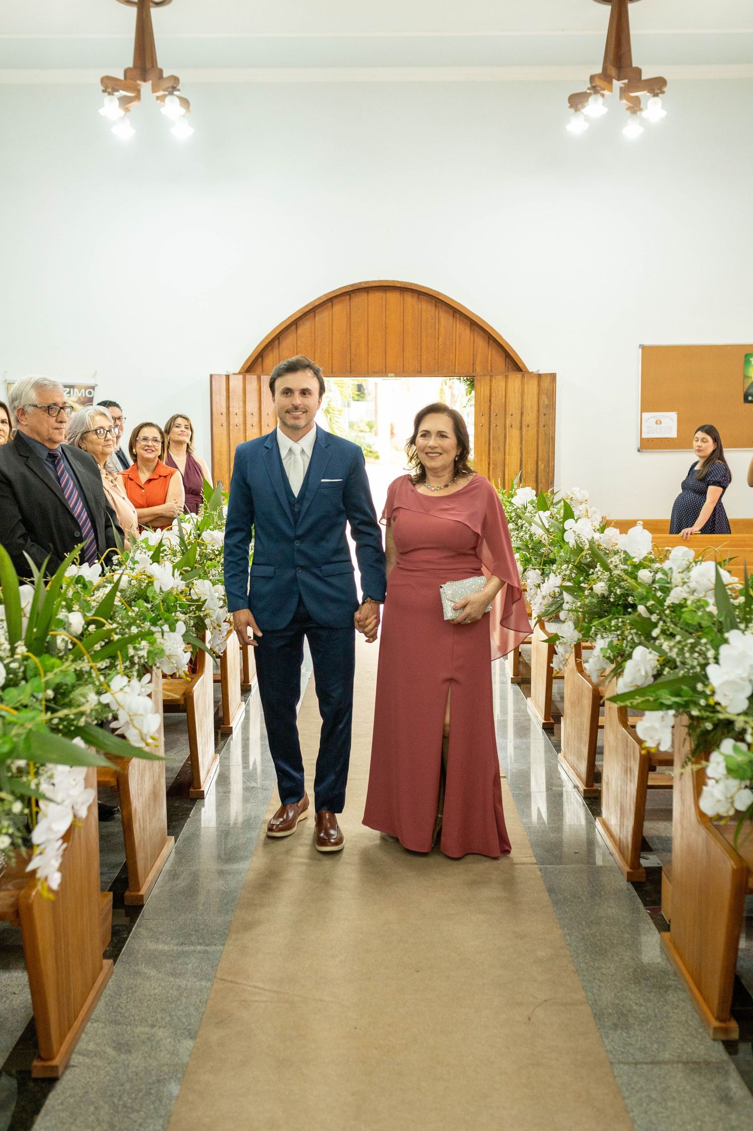 Couple walks down decorated church aisle, man in blue suit and woman in mauve dress holding hands.