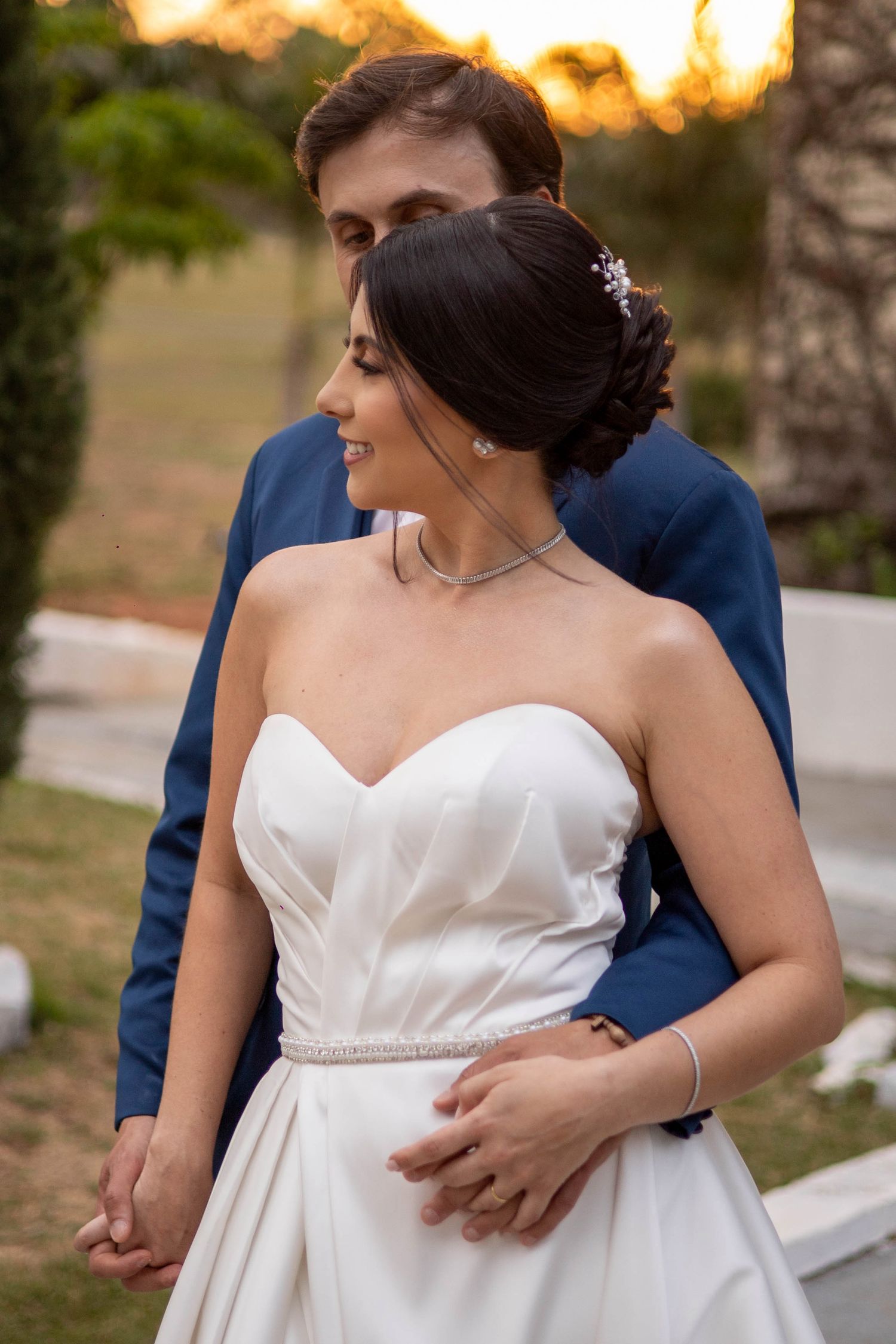 Bride in white strapless gown with groom in blue suit embracing outdoors at sunset.