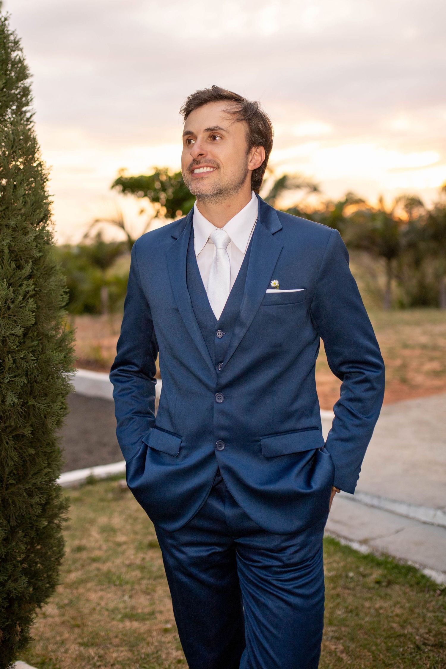 A smiling man in a navy blue three-piece suit with white tie stands outdoors at sunset with hands in pockets.