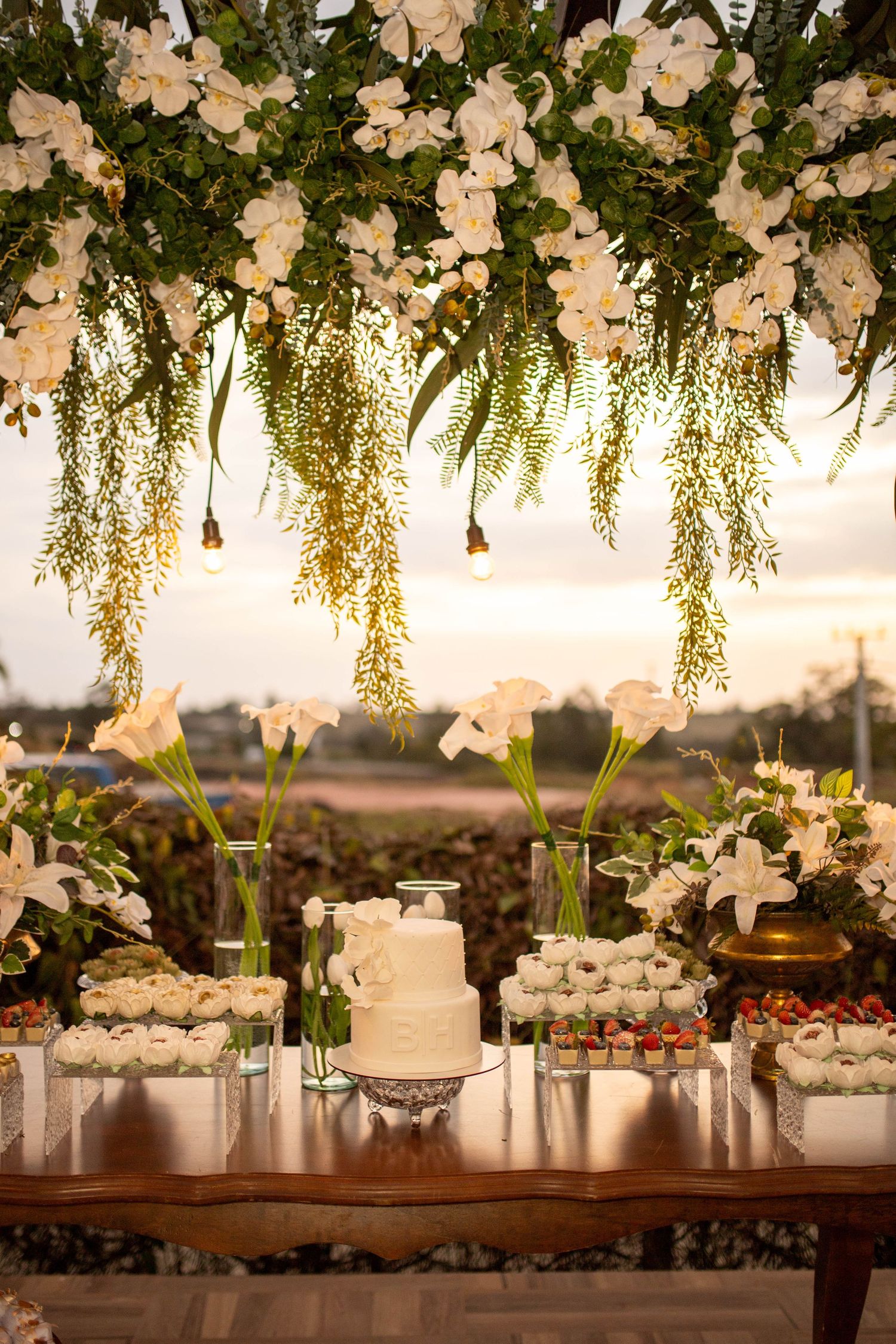 Elegant wedding dessert table with white floral arch, tiered cake, and assorted sweets at outdoor sunset reception.
