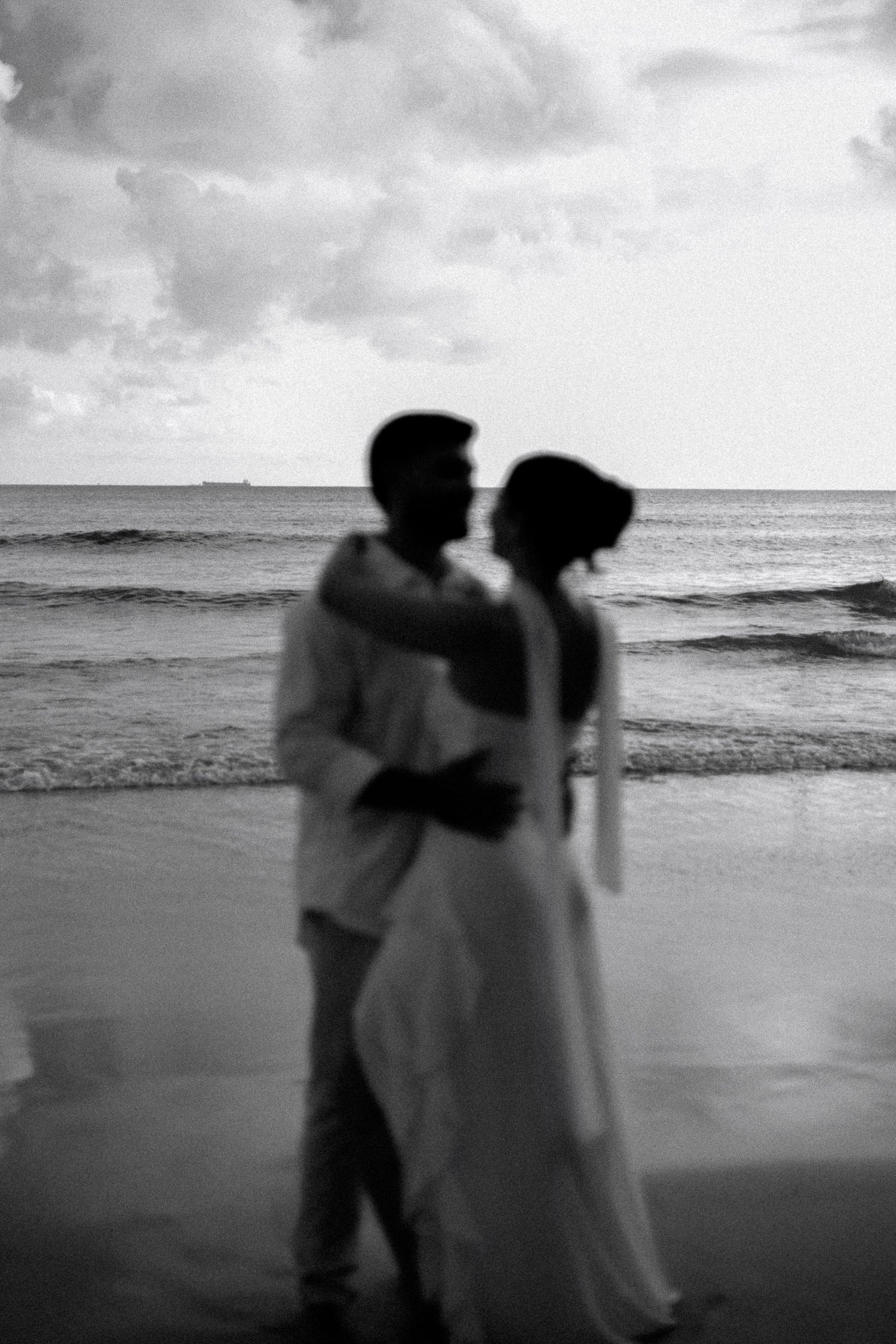Black and white photo of a couple embracing on a beach at dusk, with ocean waves and cloudy sky behind them.