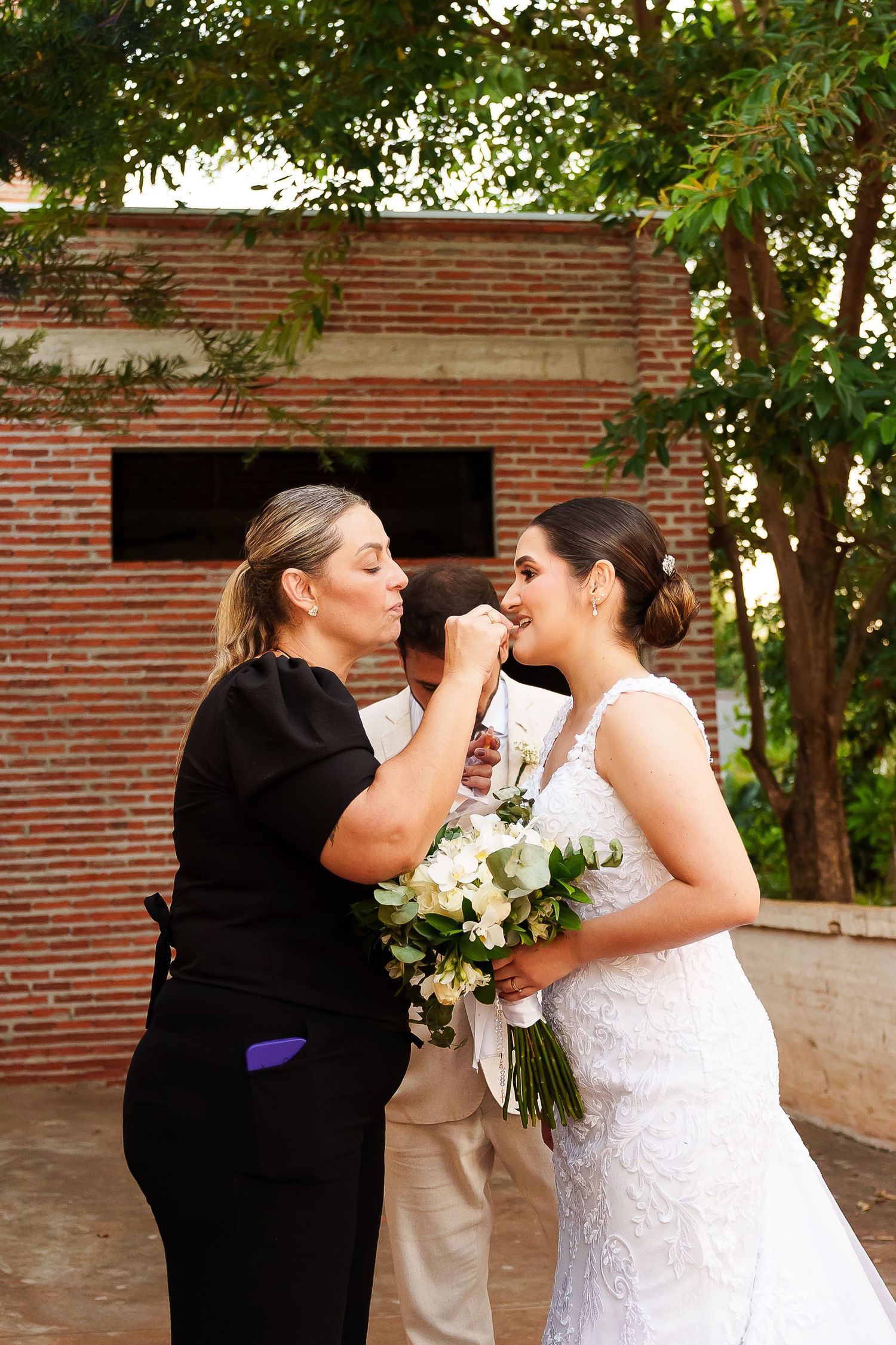 Makeup artist in black dress touching up bride in white lace gown holding floral bouquet outdoors near brick wall.