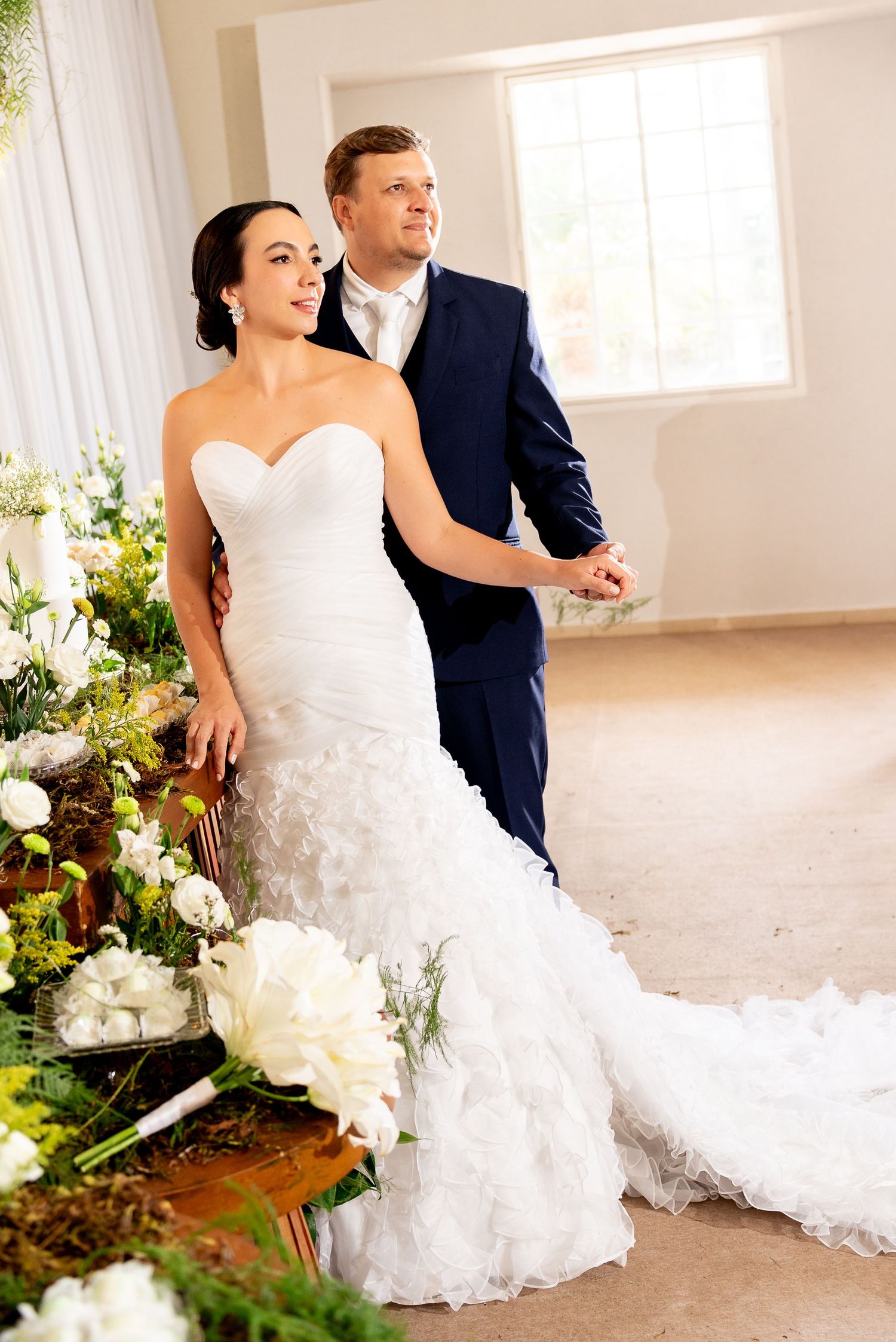 Bride in white mermaid gown and groom in navy suit pose together near floral arrangements at elegant wedding venue.