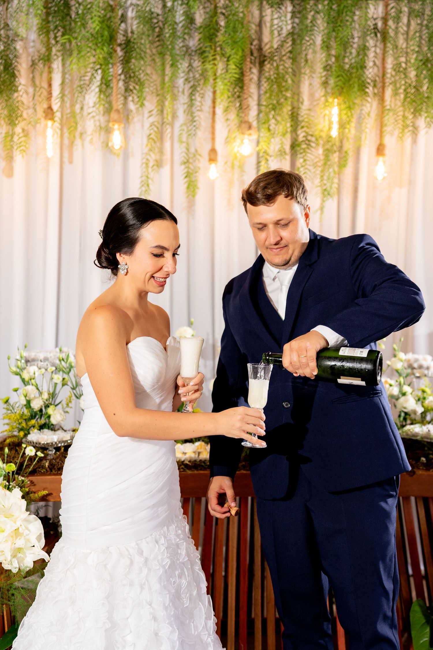 Bride and groom pouring champagne at wedding reception under hanging greenery and Edison bulb decorations.