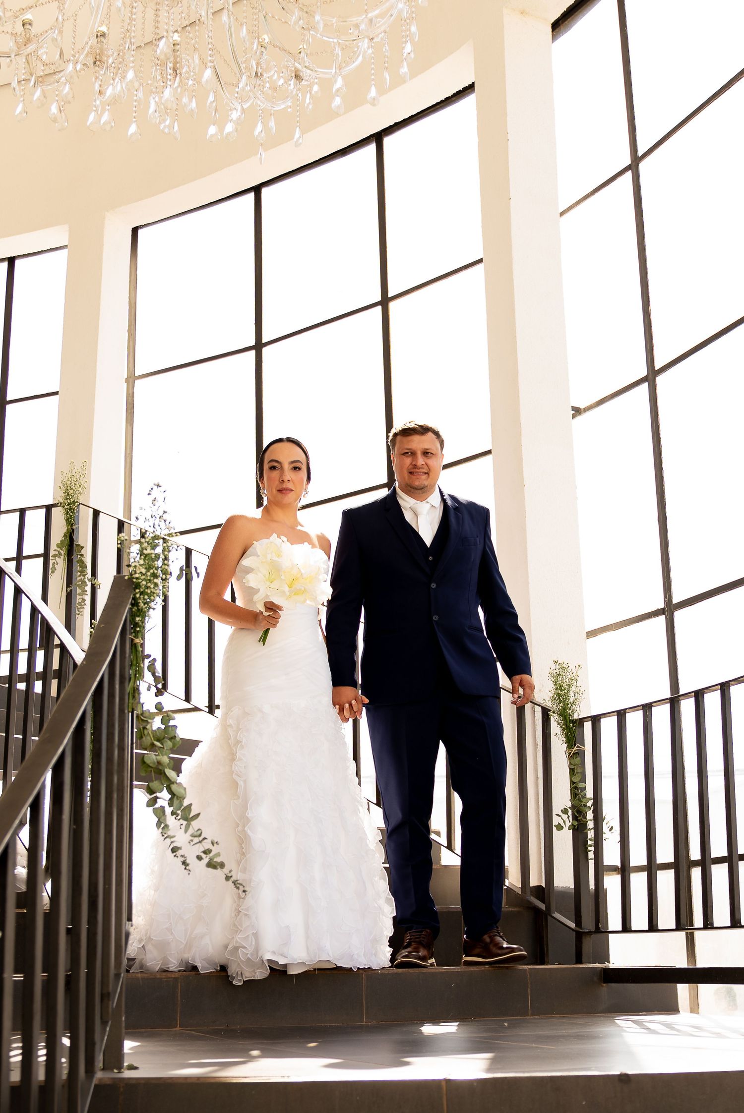 Bride in white ruffled gown and groom in navy suit descend elegant staircase with large windows and chandelier.