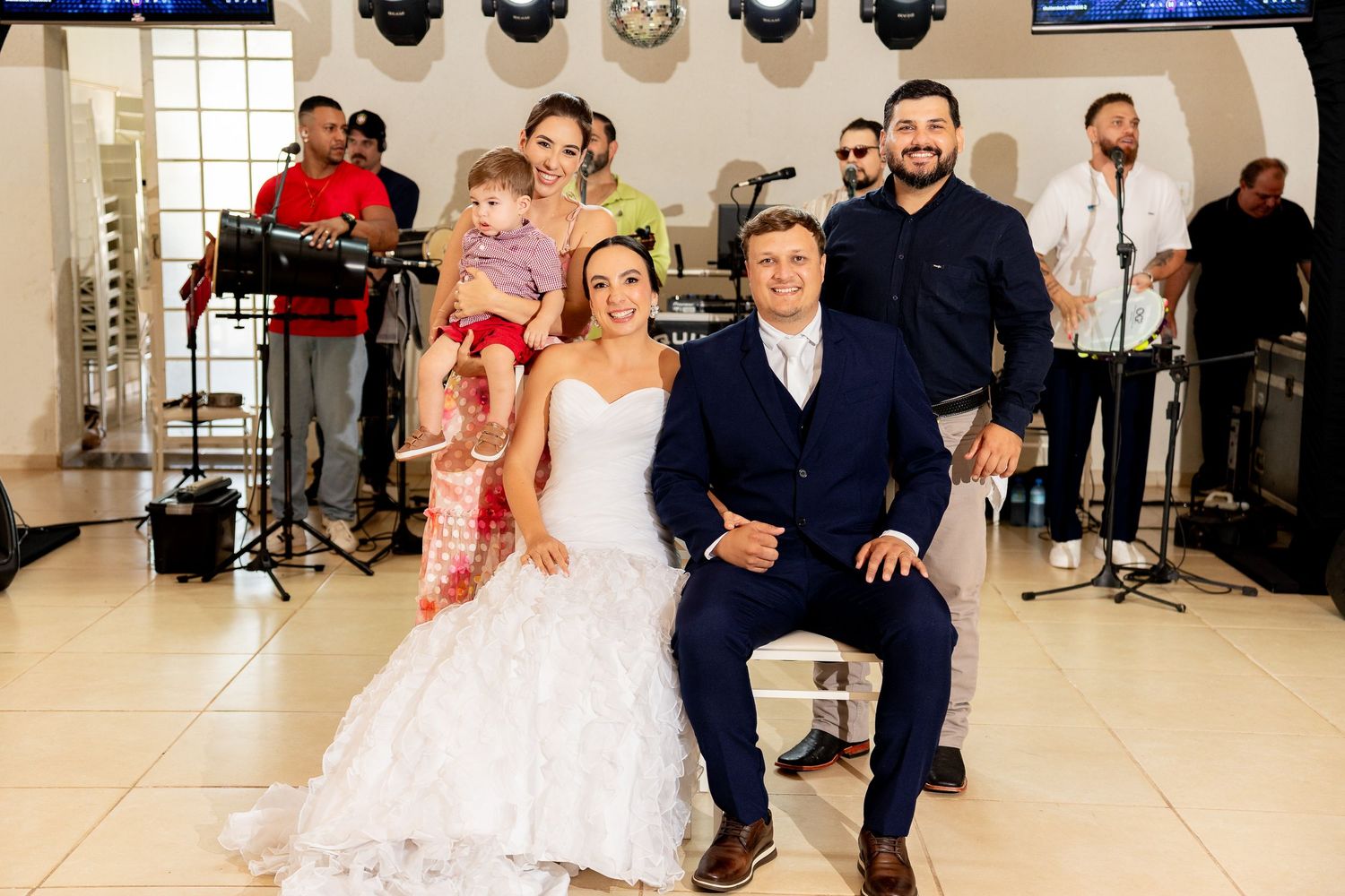 Bride and groom pose with wedding guests and live band performing in background at elegant reception venue.