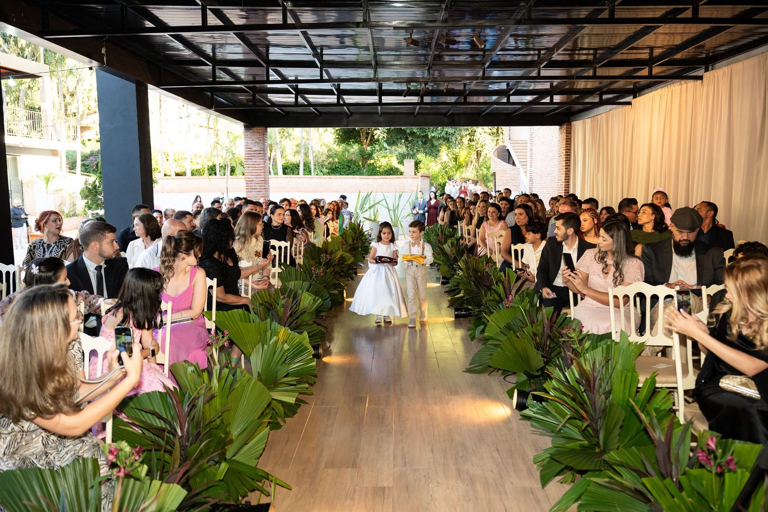 Elegant outdoor wedding ceremony with guests seated along a lush tropical aisle as flower girls walk toward the altar.