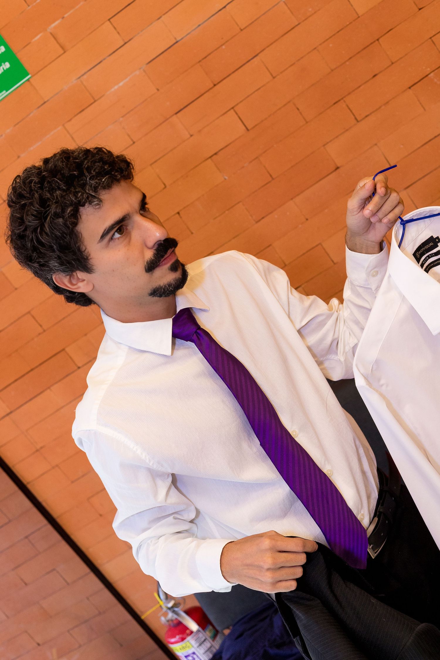A man in a white shirt and purple tie writes on a whiteboard against an orange brick wall.