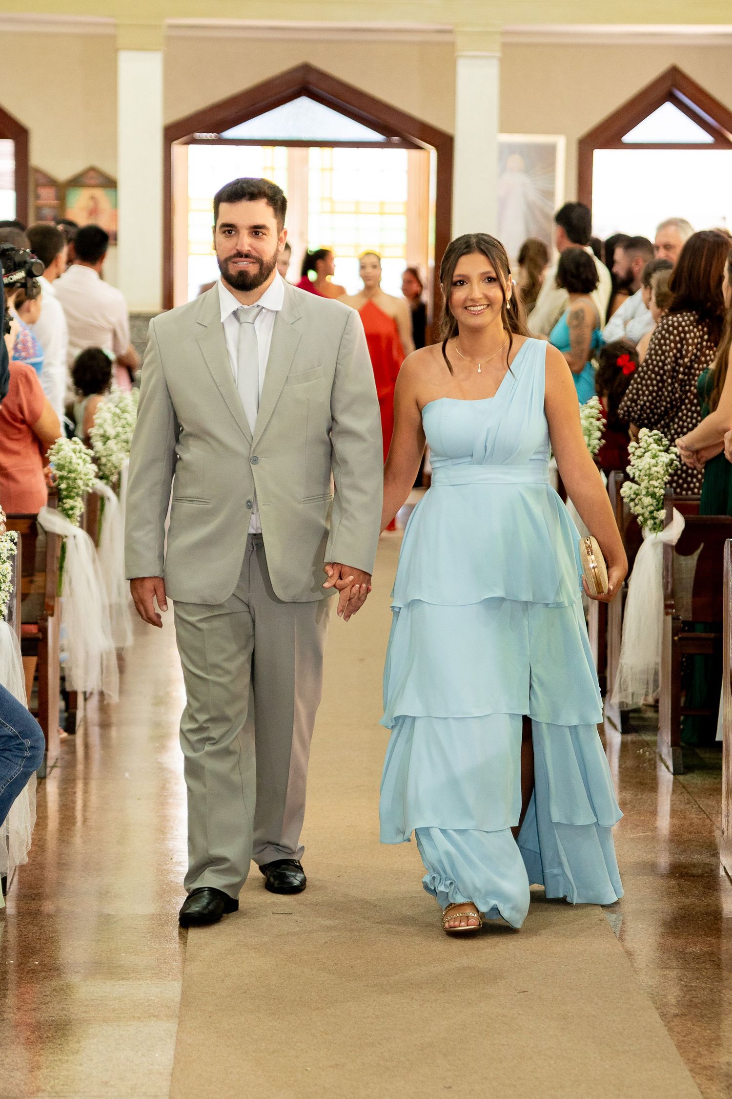 A couple walks down a church aisle, man in gray suit and woman in light blue tiered gown, at a wedding ceremony.