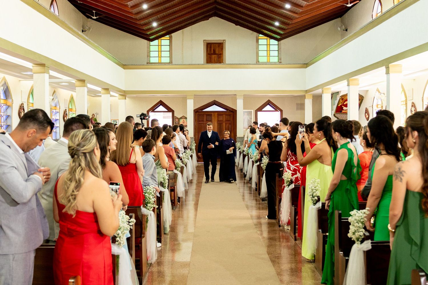 Wedding guests stand in church pews as groom walks down aisle flanked by bridesmaids in red and green.