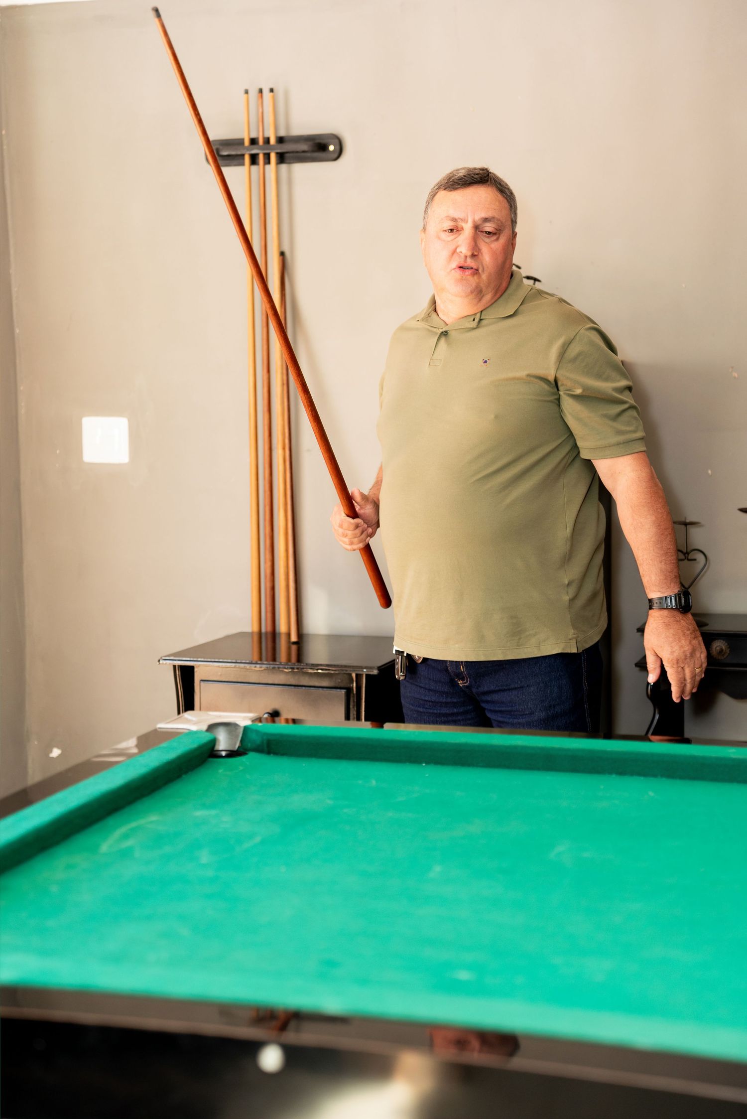 Middle-aged man in green polo shirt holding a billiard cue stick beside a green pool table indoors.