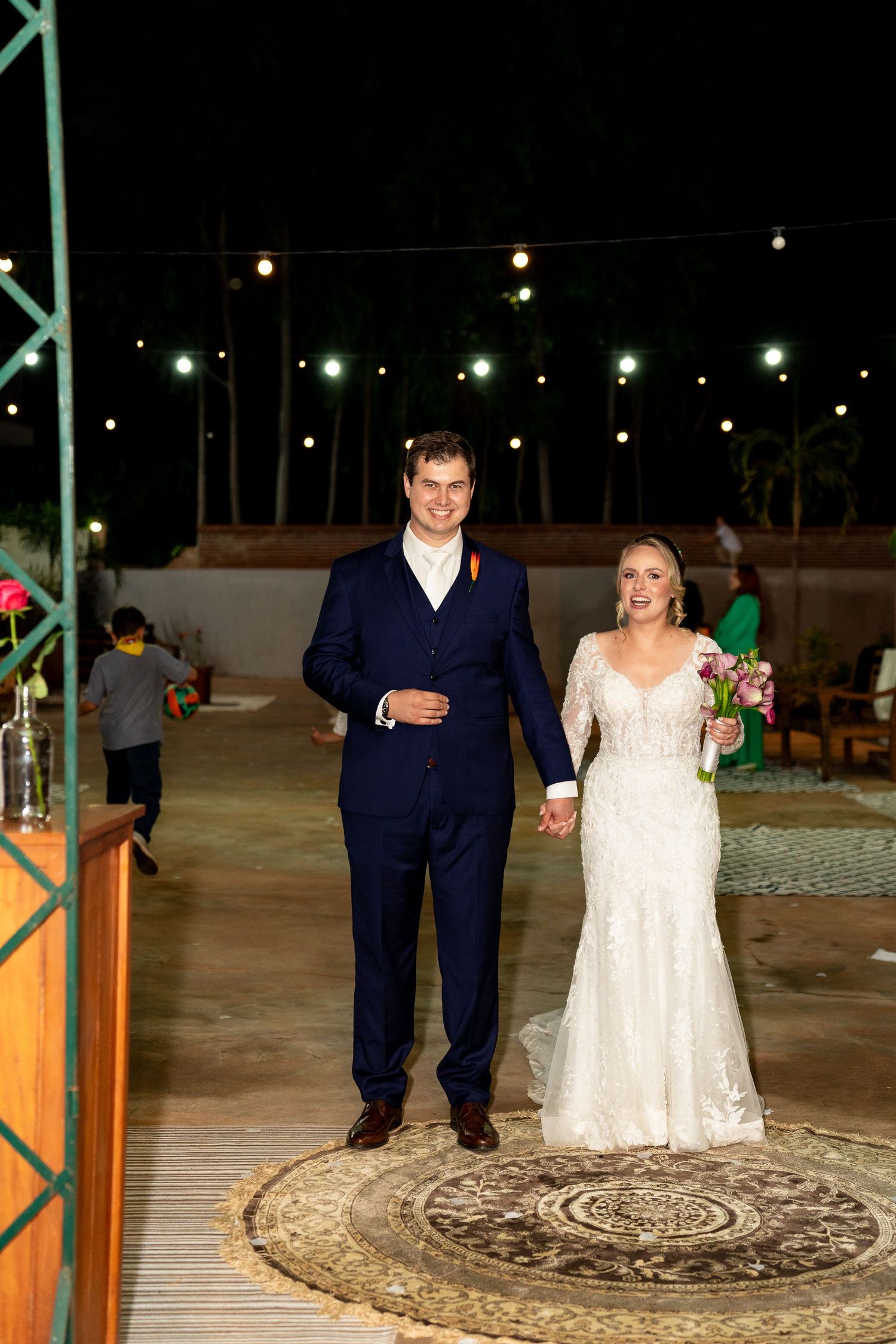Bride in lace gown and groom in navy suit hold hands smiling at outdoor night wedding with string lights.
