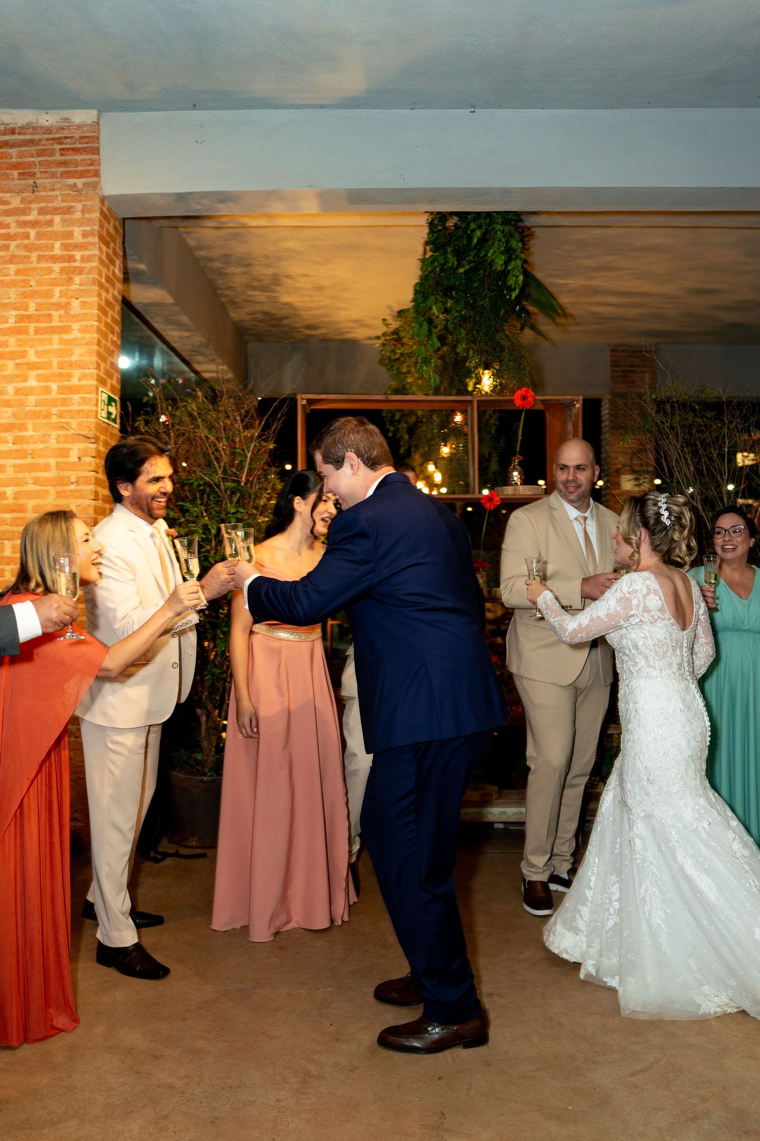 Bride and groom share a toast with guests at elegant indoor wedding reception with brick walls and greenery.