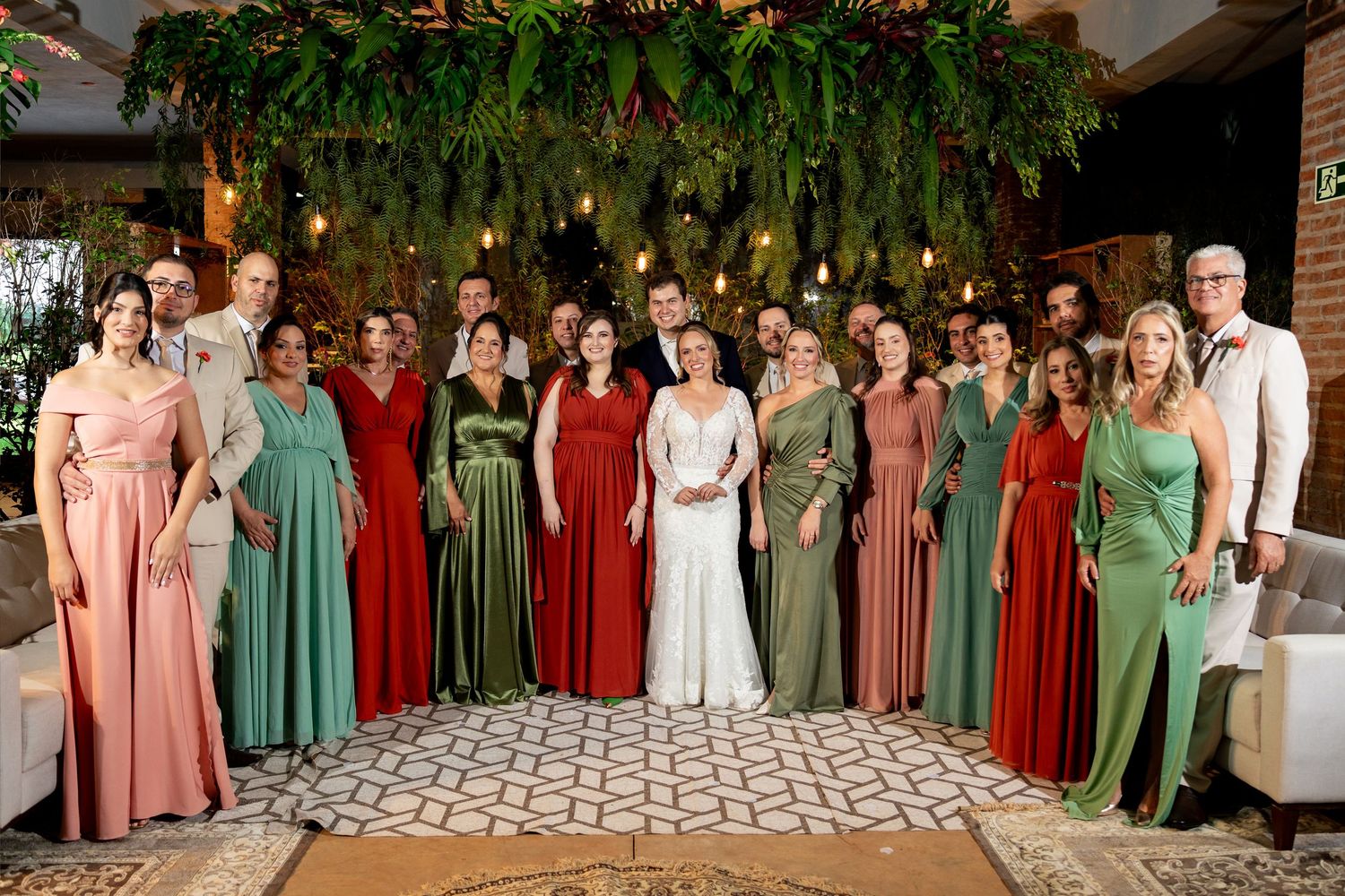 Wedding party poses together in red and green gowns with bride and groom centered under lush floral greenery backdrop.