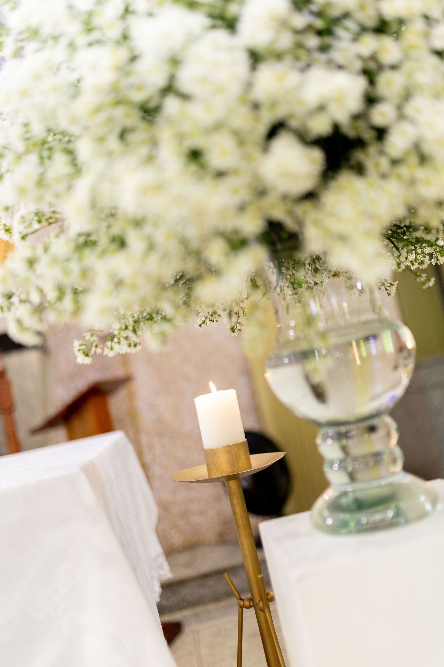 Elegant wedding altar with white floral arrangement, lit candle on gold holder, and crystal glass on white draped table.