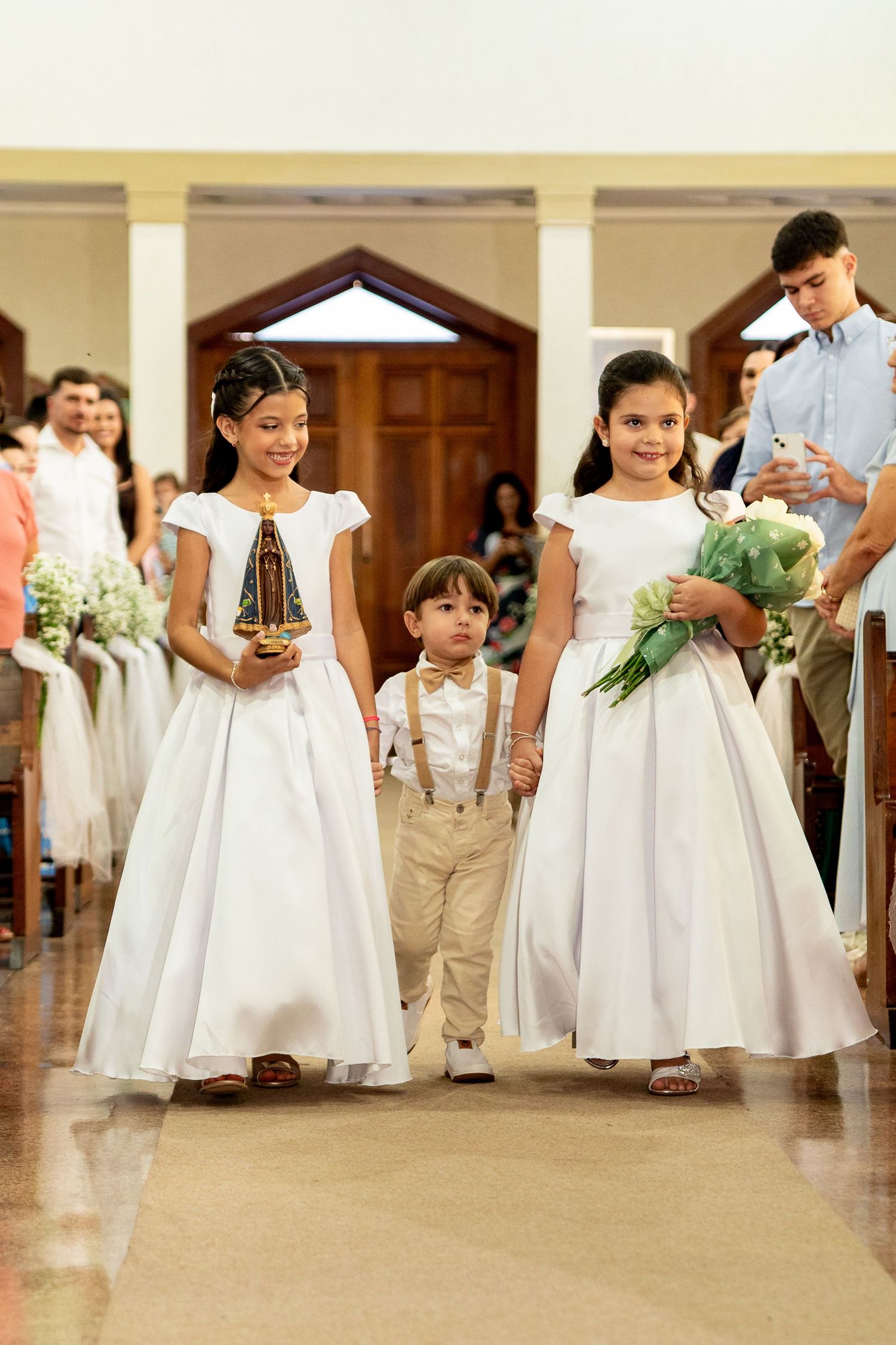 Two flower girls in white dresses and a ring bearer walk down a church aisle during a wedding ceremony.