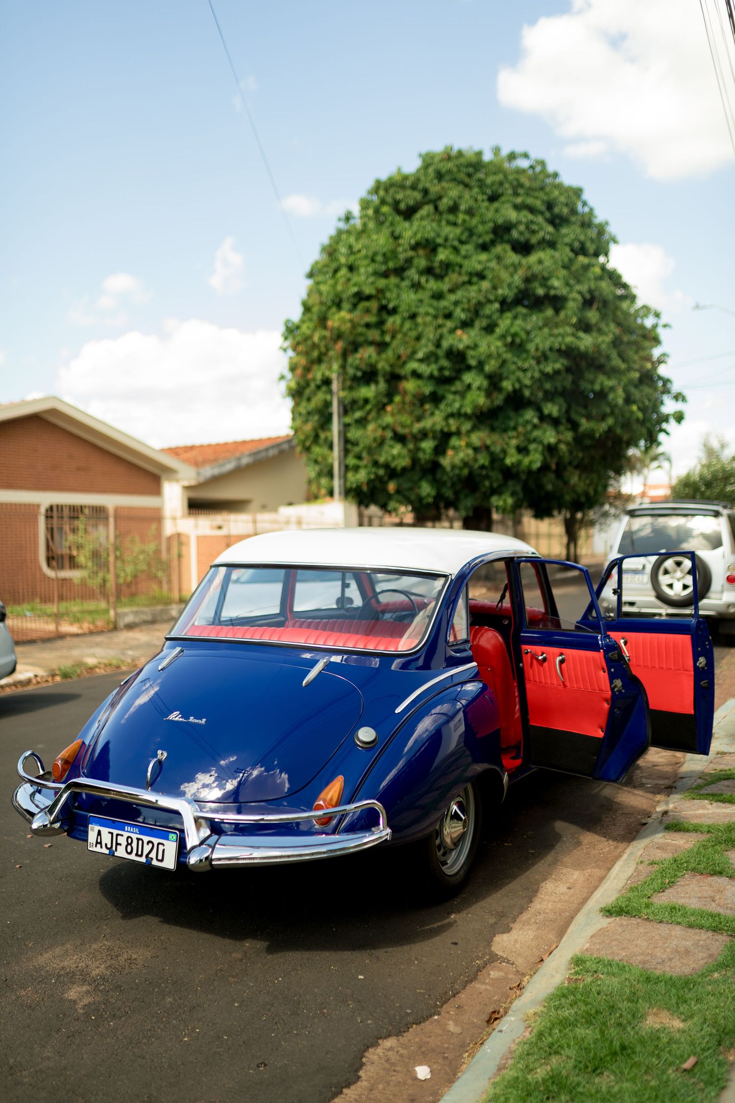 Vintage blue classic car with red interior and open door parked on a suburban street with green trees in background.