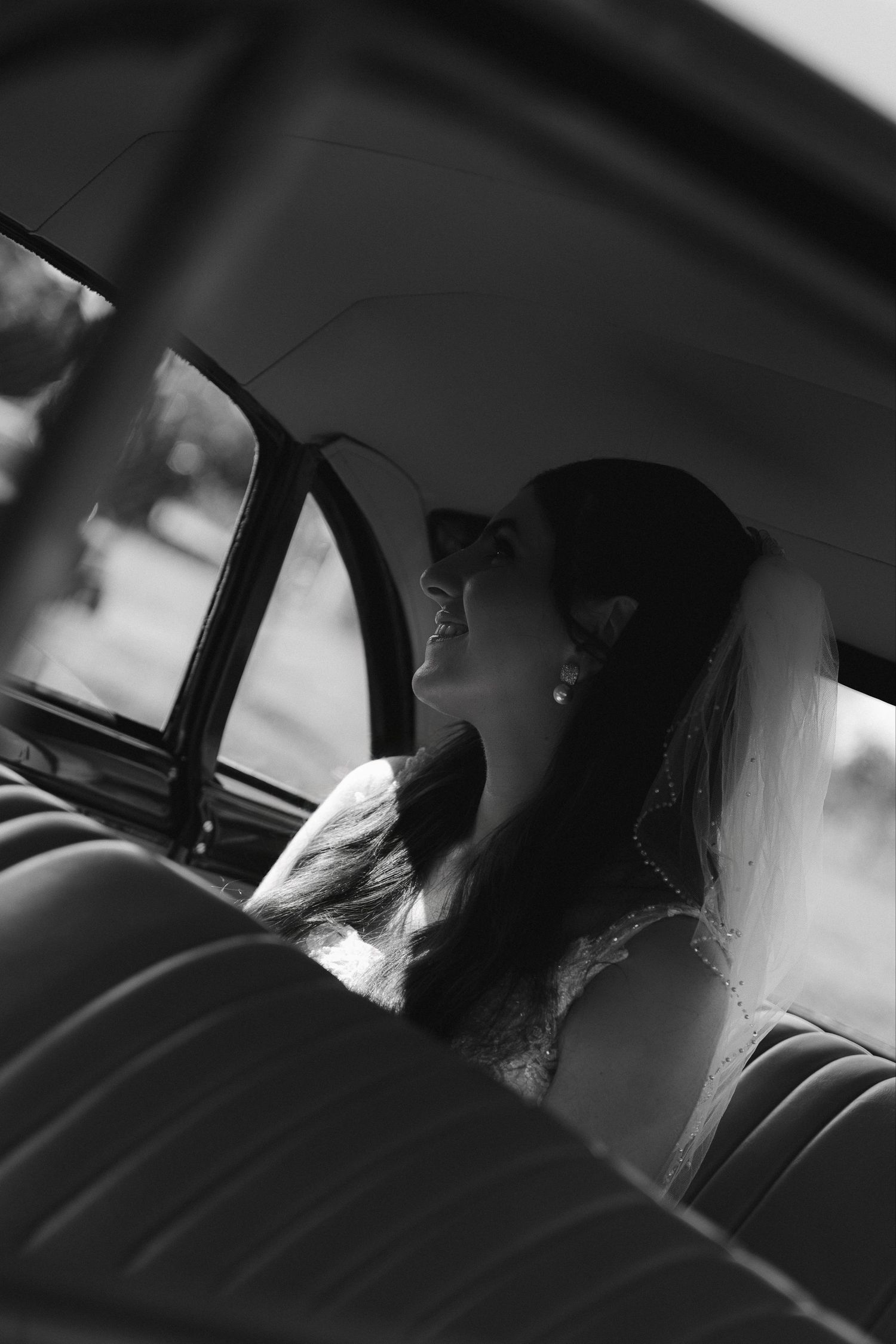 Black and white photo of a smiling bride in a veil sitting in a vintage car backseat on her wedding day.