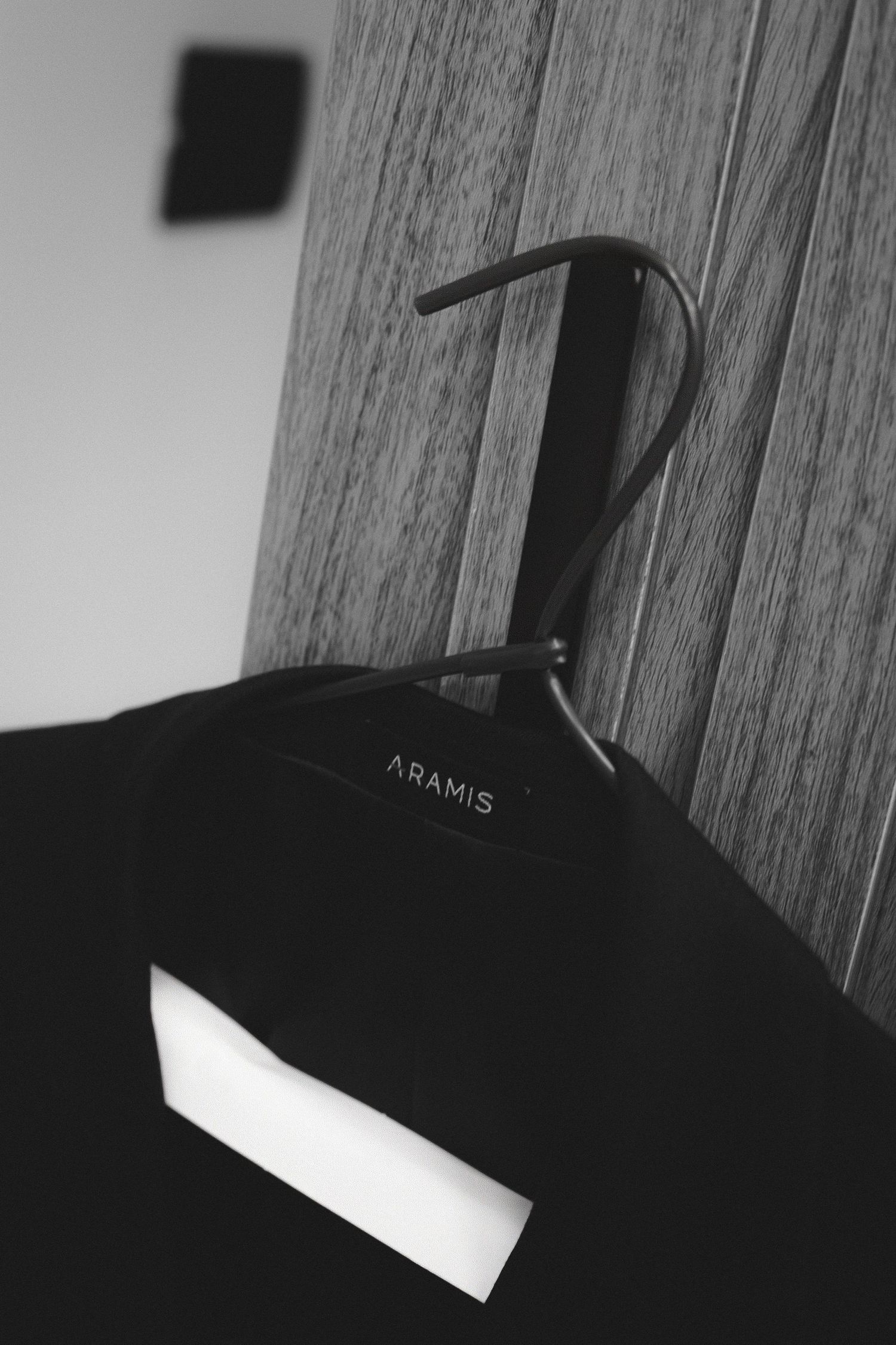 Black and white photo of an Aramis branded clothing hanger with a dark garment against a wooden wall background.