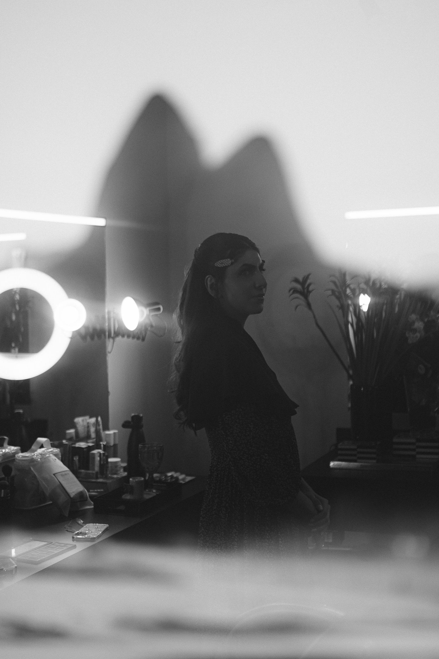 Black and white photo of a woman in a dressing room with ring light and shadow on wall.
