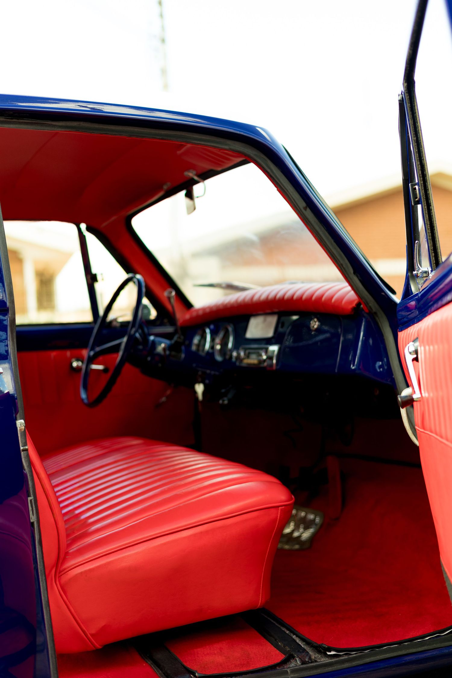 Vintage car interior with vibrant red leather seats and dashboard, black steering wheel, and blue exterior visible through open door.