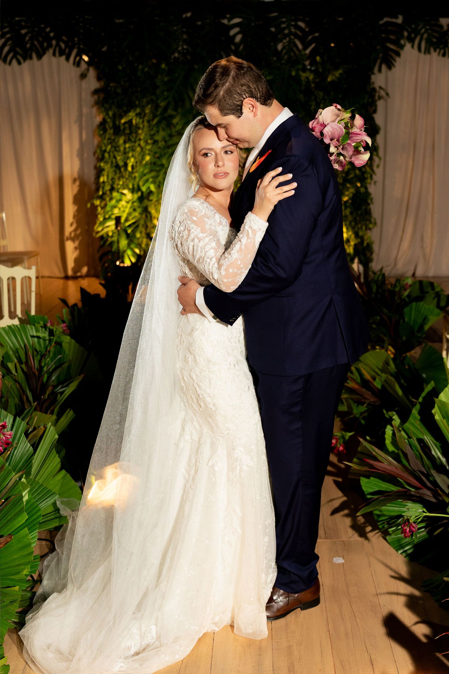 Bride in lace gown and veil embraces groom in navy suit, holding pink bouquet, surrounded by tropical greenery.
