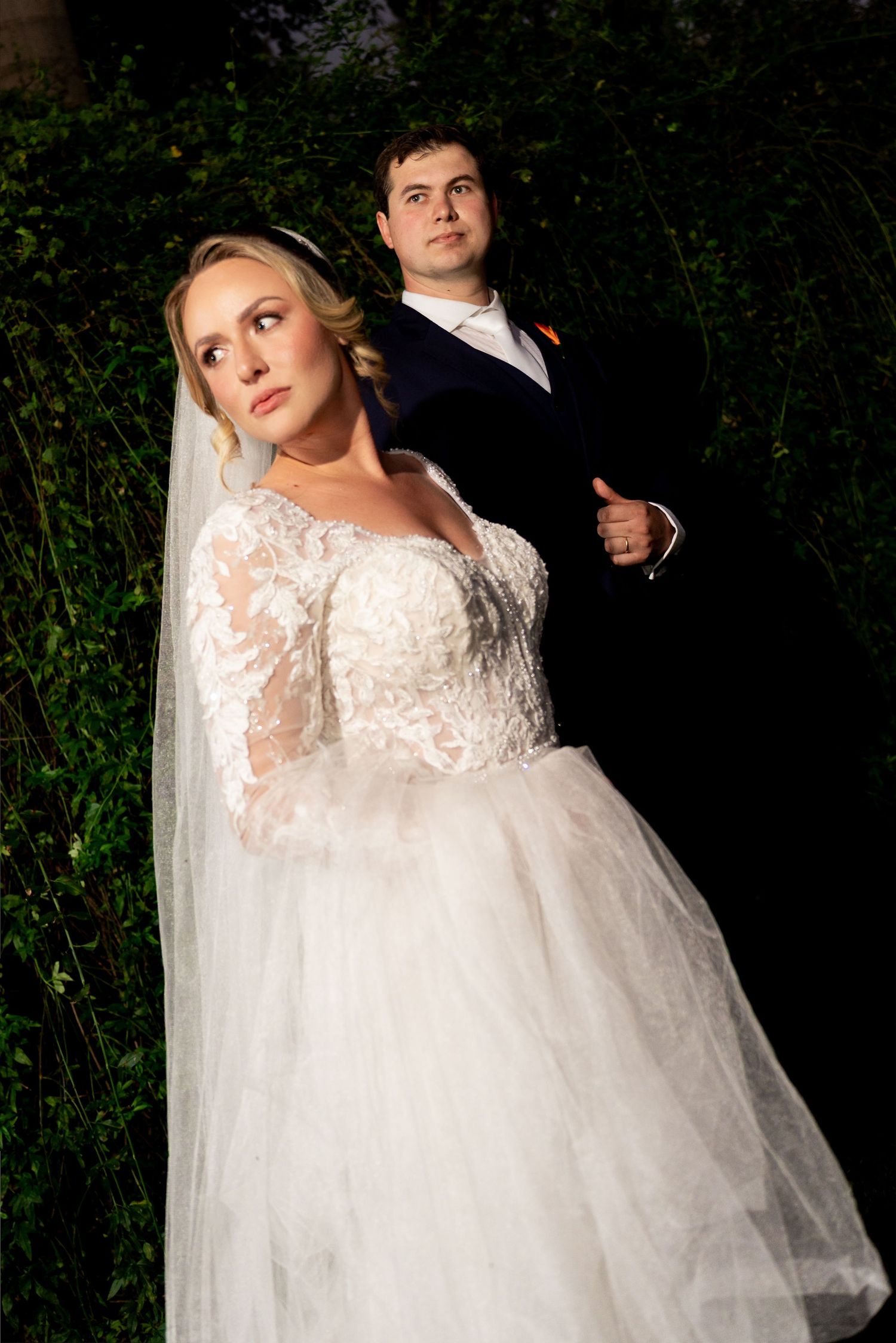 Bride in lace ballgown with veil stands before groom in black tuxedo amid lush greenery at elegant wedding.