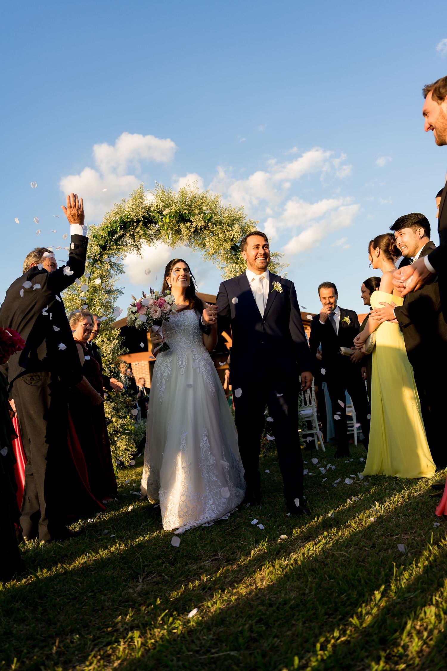 Bride and groom walk through cheering guests under floral arch at outdoor wedding ceremony on sunny day.