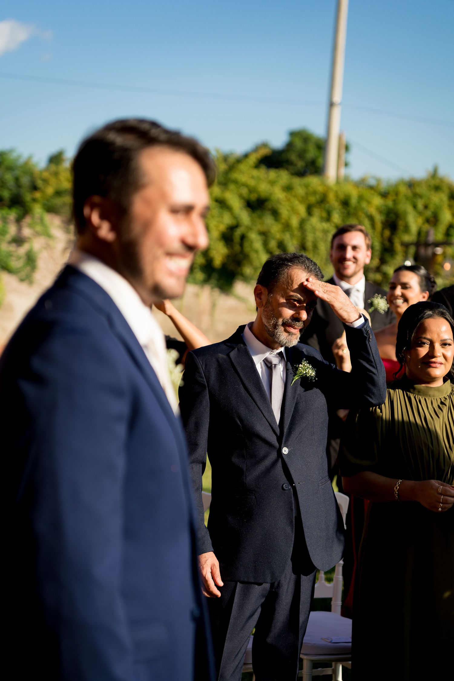 Emotional father wiping tears at outdoor wedding ceremony while groom stands in foreground in navy suit.