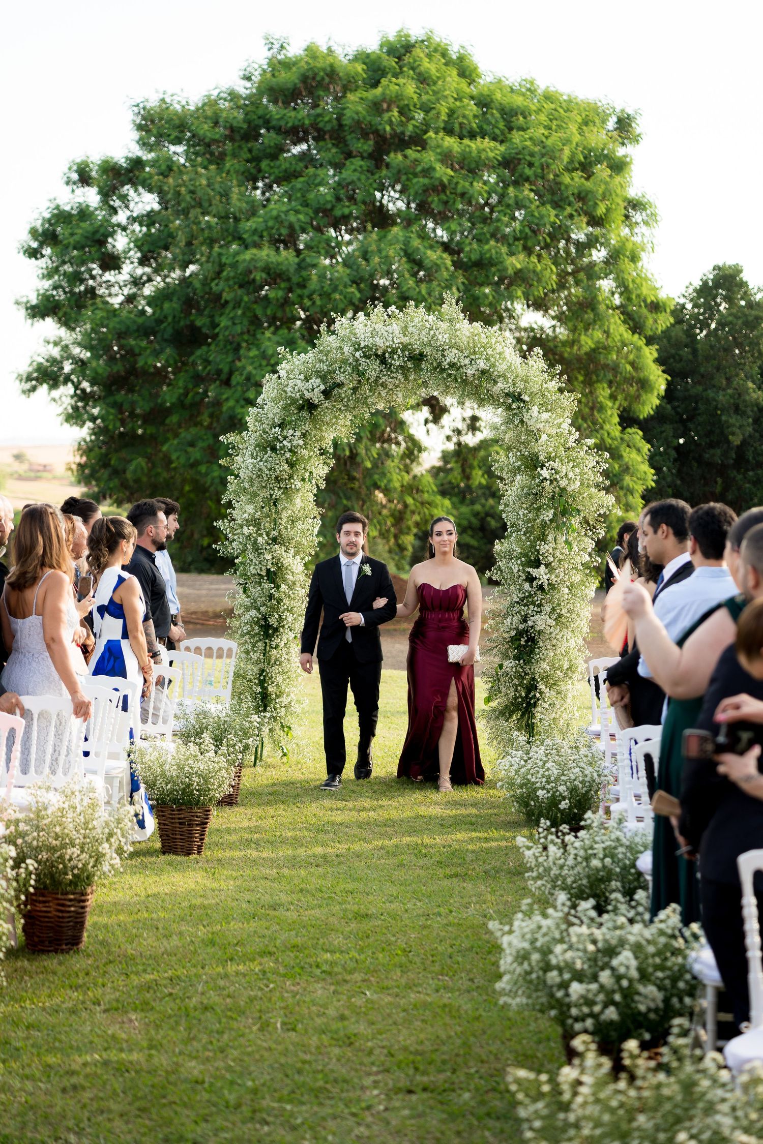 Couple walks down floral arch aisle at outdoor wedding ceremony on lush green lawn with white chairs and baby's breath decor.