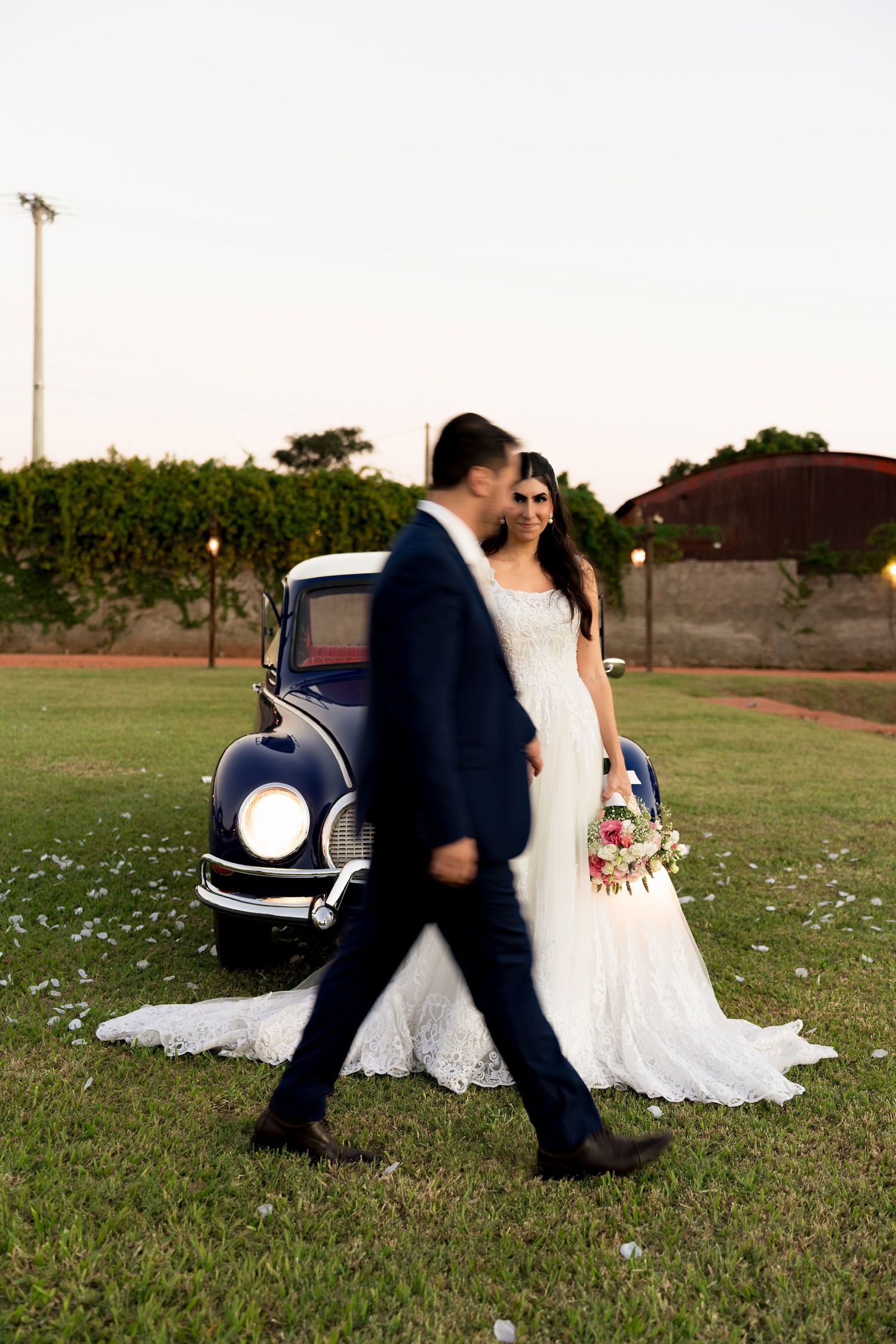 Bride and groom walking together in front of a vintage blue car on a green lawn at sunset.