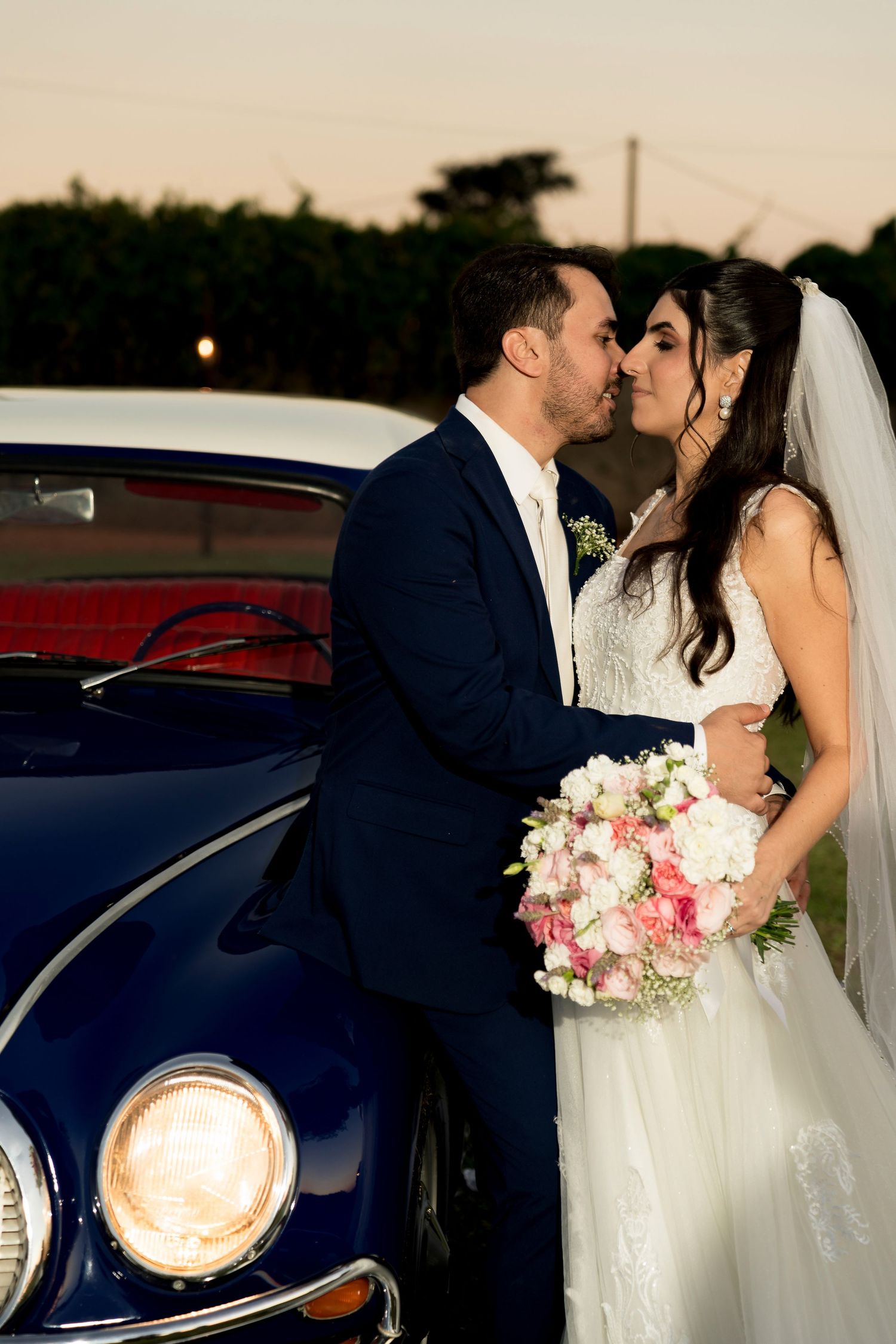 Bride and groom sharing a romantic kiss beside a classic blue car at sunset, bride holding a pink and white bouquet.