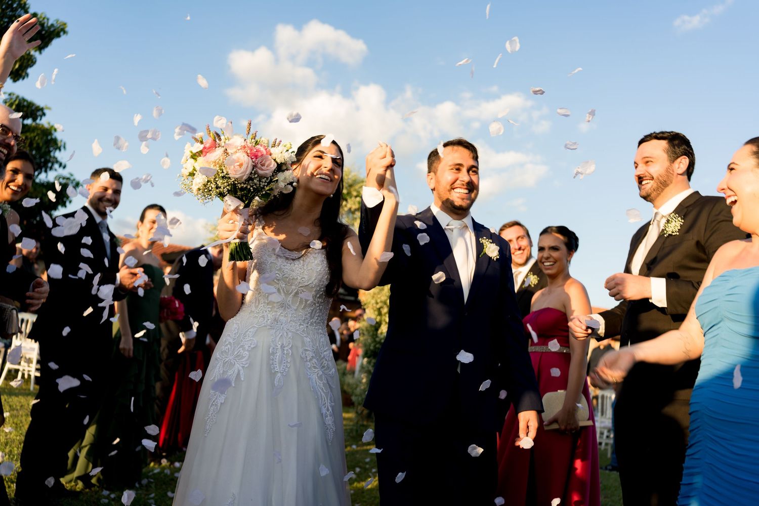 Newlywed couple walks through confetti shower, bride holding bouquet, guests celebrating outdoors at wedding ceremony.
