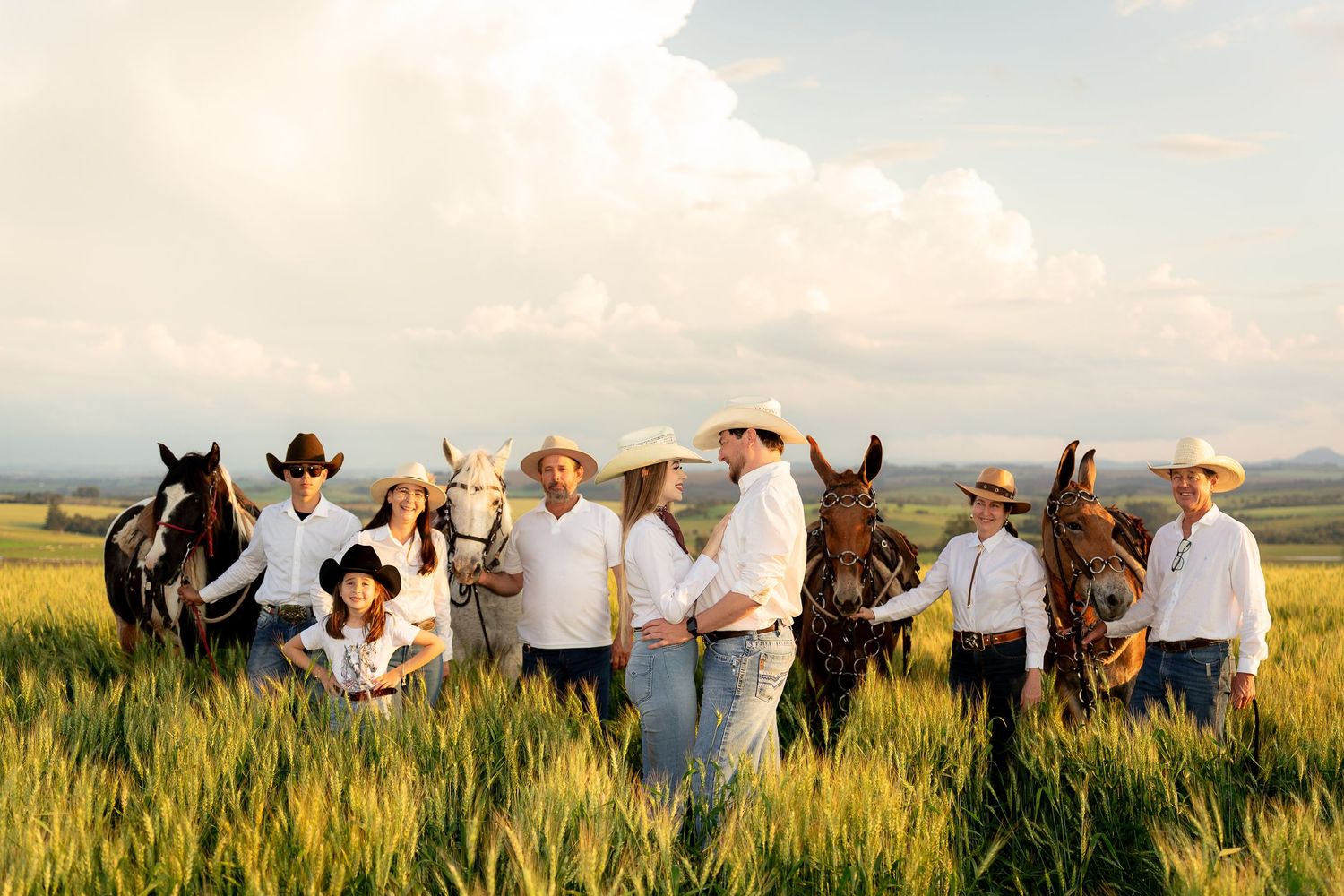 A family in white shirts and cowboy hats poses with horses in a lush green field under a cloudy sky.