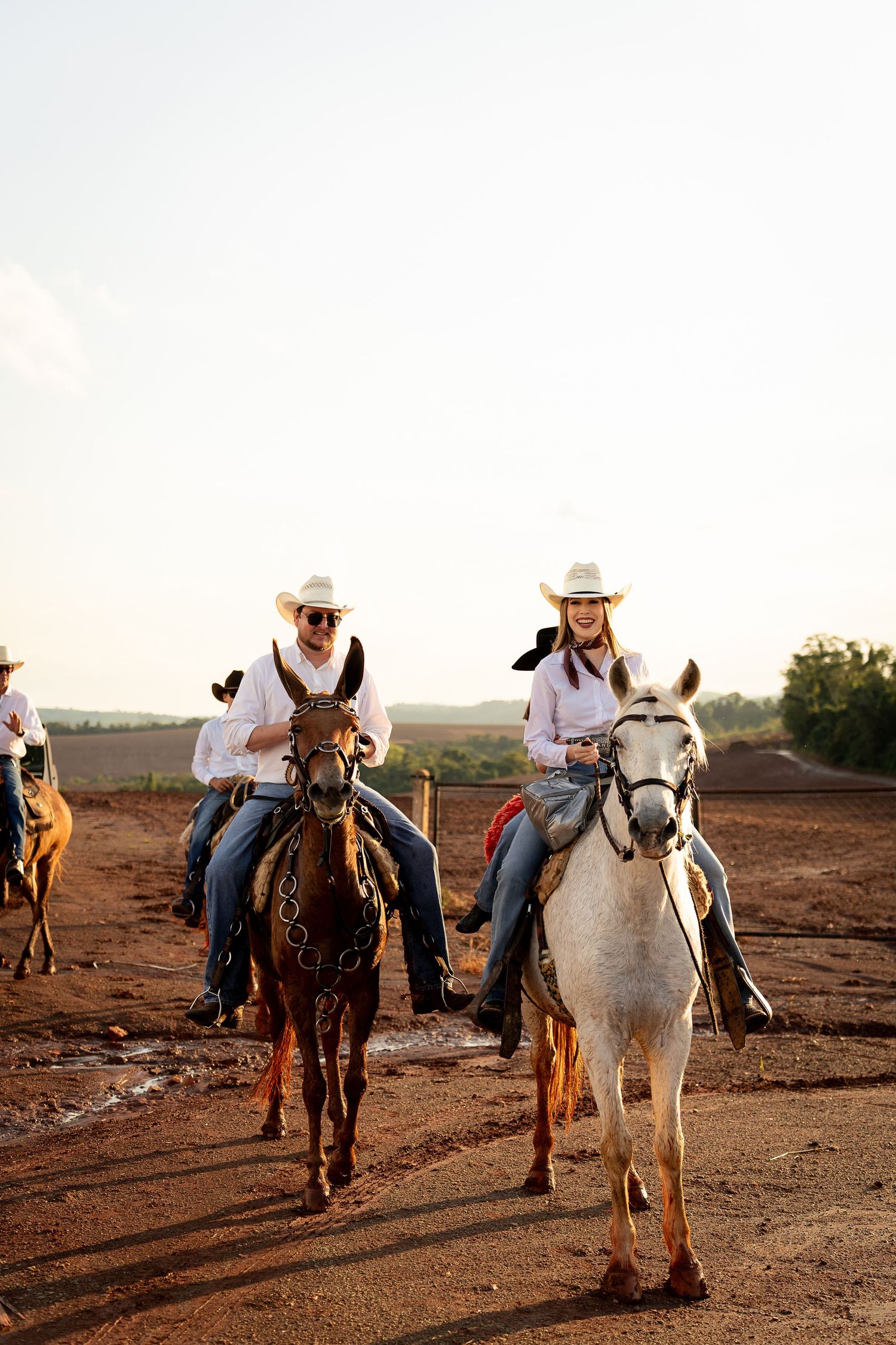 Two cowboys riding horses on a dirt trail at sunset, wearing white hats and shirts in a rural landscape.