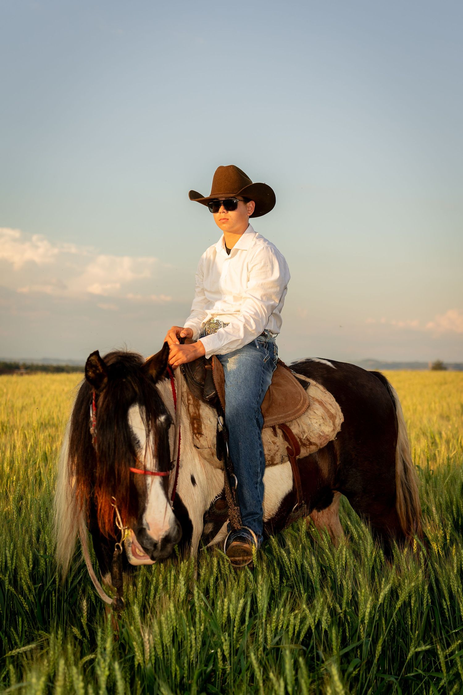 A young cowboy in a white shirt, jeans, and brown hat rides a paint horse through a green field.